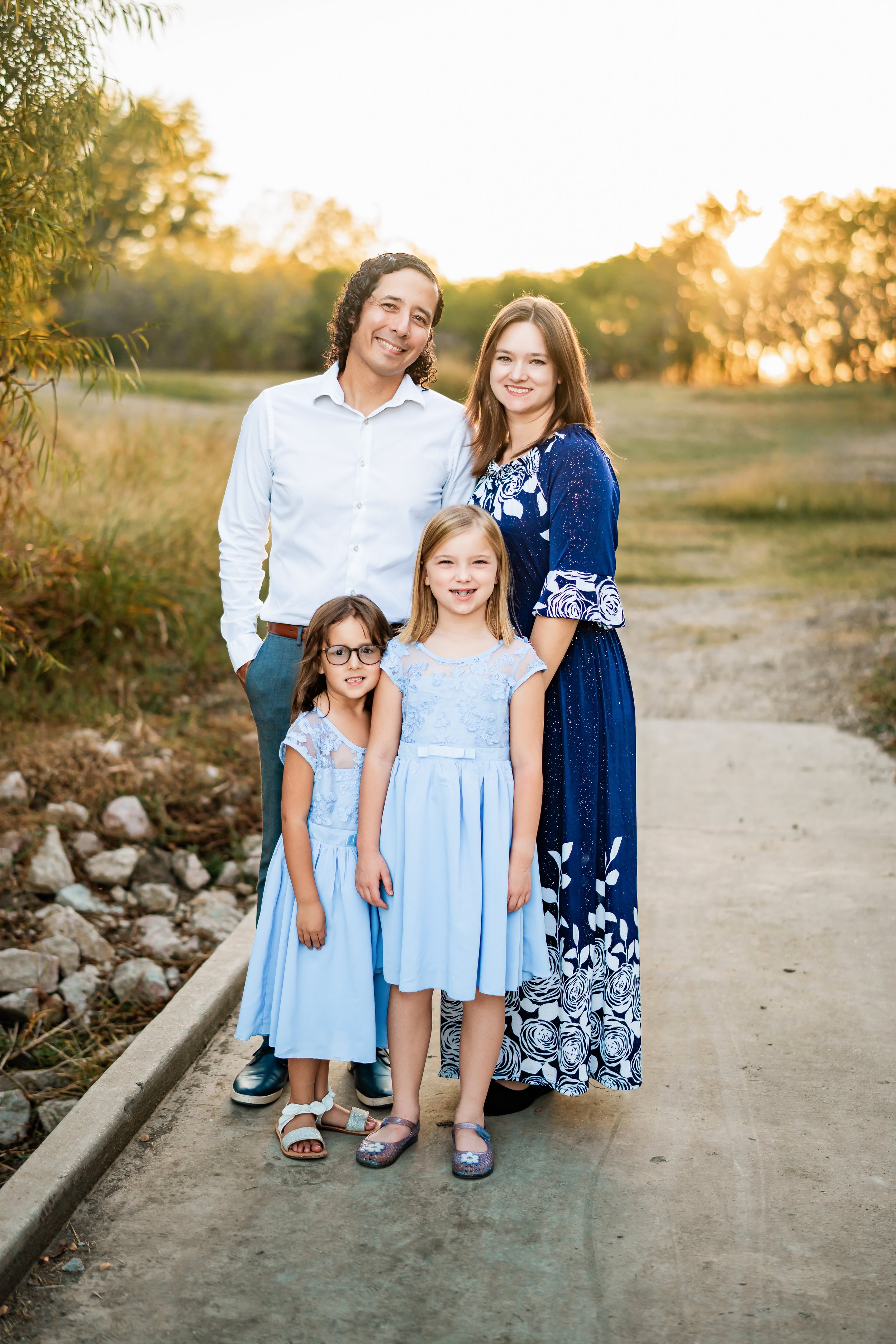 A family of five standing outdoors on a dirt path with trees and sunset in the background, smiling at the camera. The family includes two adult women, one adult man, and two young girls, all dressed in blue and white clothing.