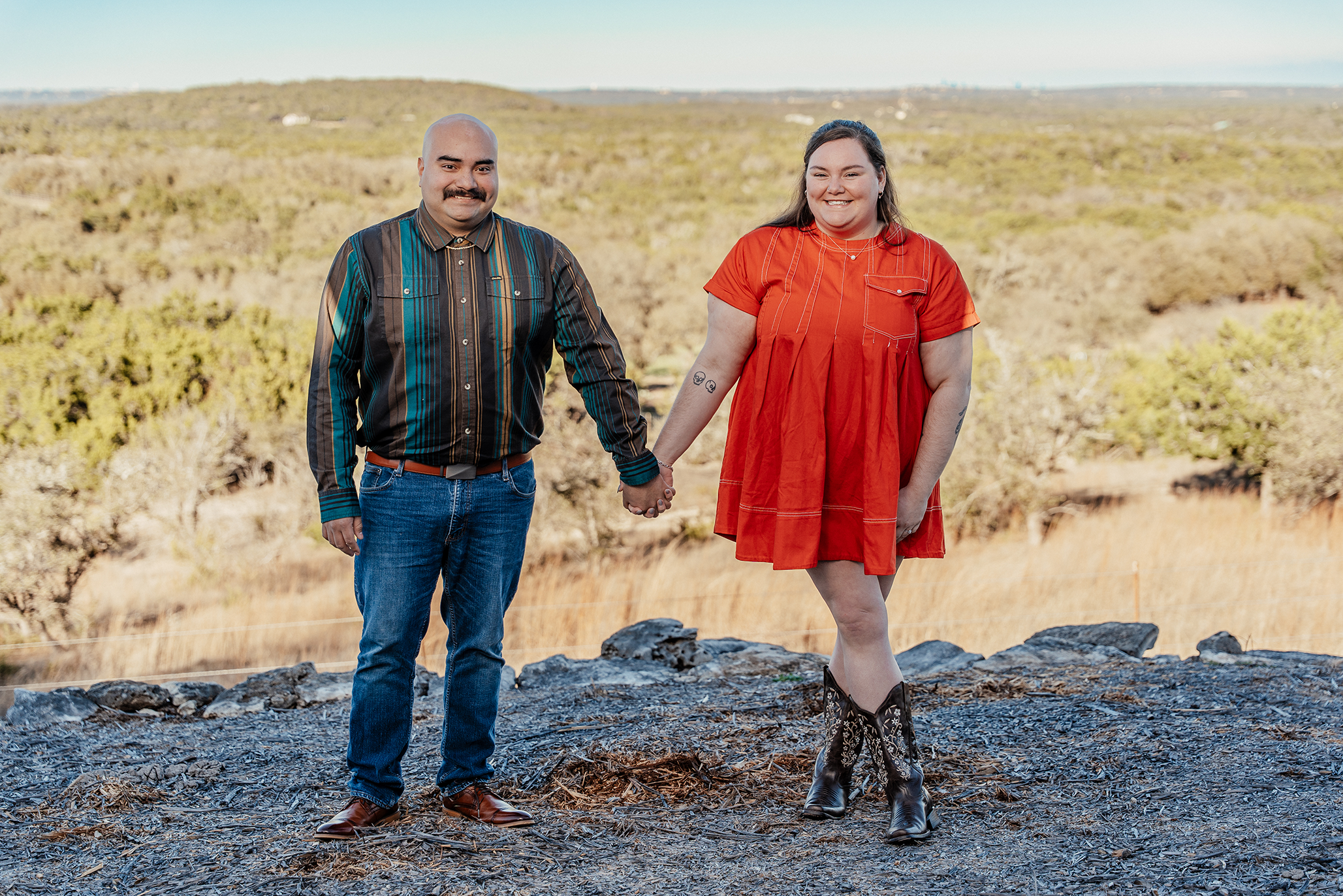 A couple holding hands and smiling outdoors on a hilltop with a scenic landscape of trees and open land in the background.