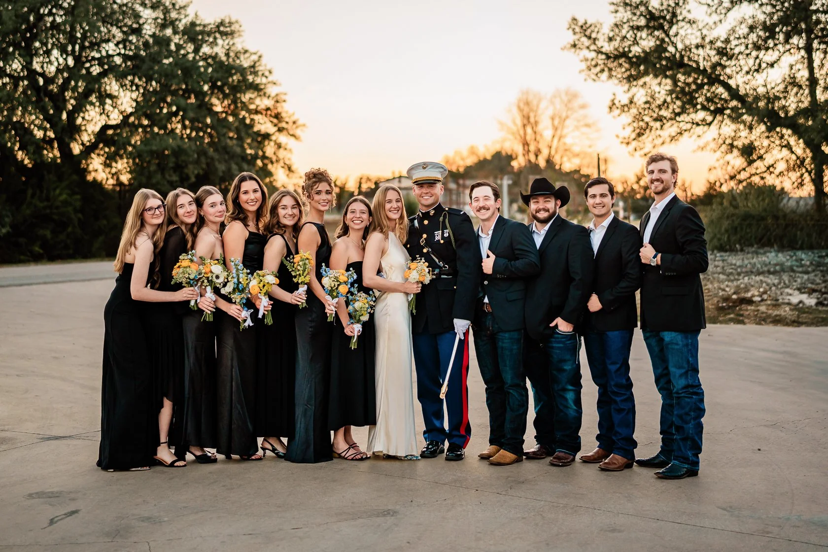 Group of people at a wedding, dressed in formal attire, standing outdoors at sunset, smiling for the camera.