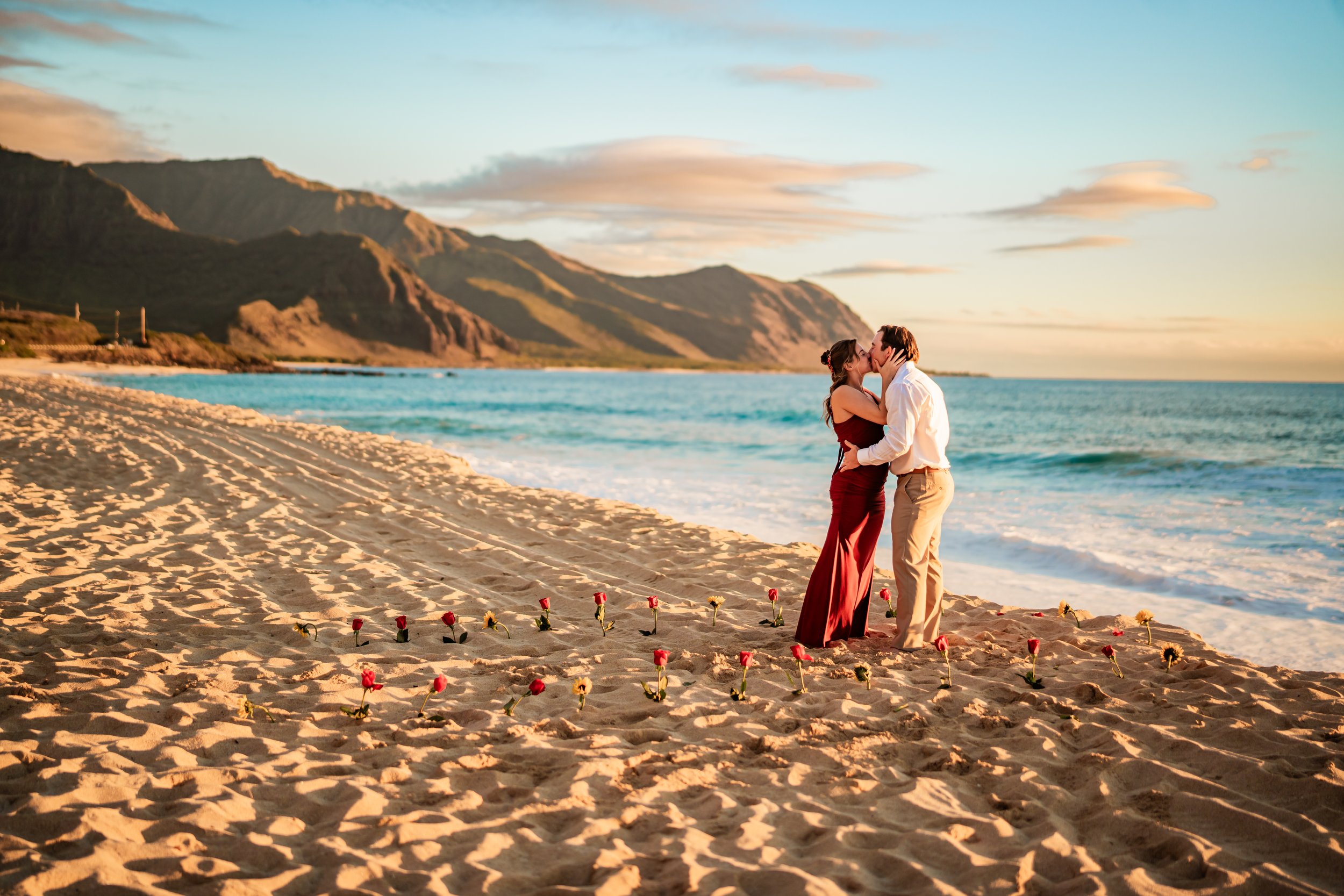 A couple standing on a sandy beach at sunset, with mountains in the background, sharing a kiss surrounded by small flowers laid in a line on the sand.