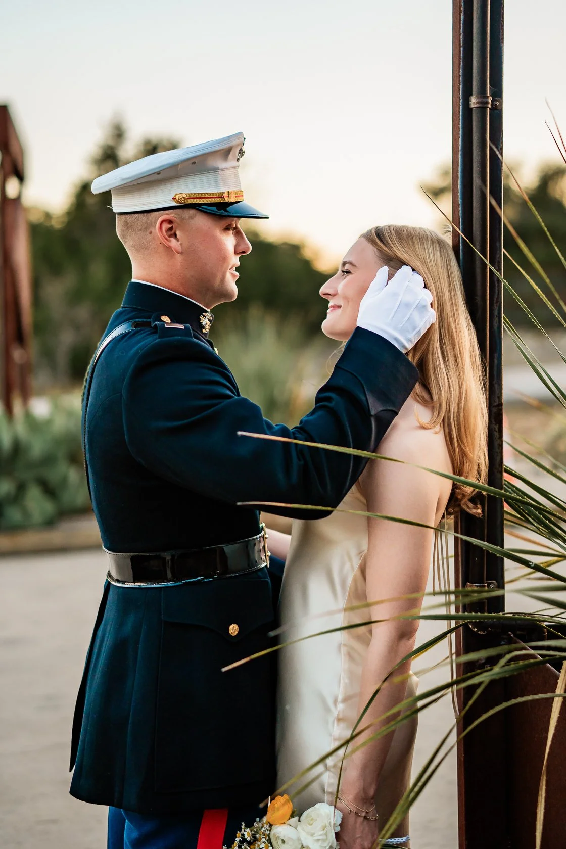 A military officer in uniform gently holds a woman's face, standing outdoors at sunset, with the woman smiling and holding a small bouquet of white flowers.