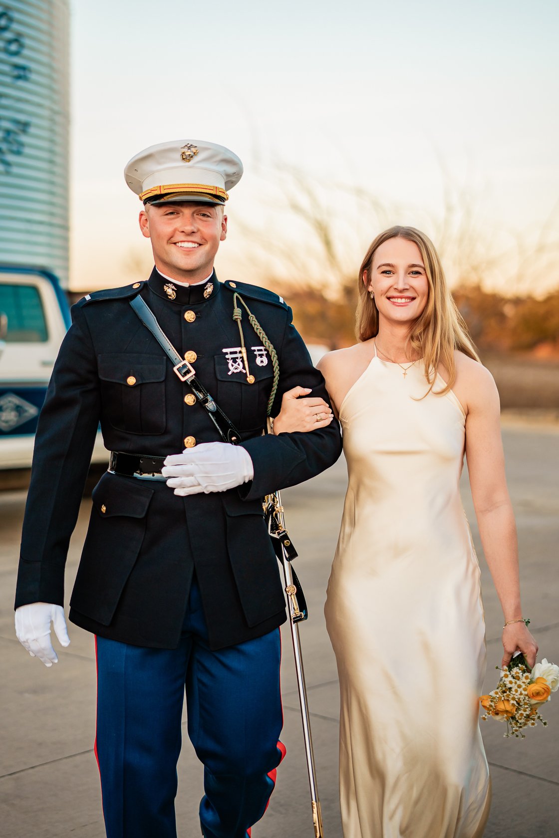 A man in a military dress uniform, smiling, holding a woman’s arm. The woman, wearing a light-colored satin dress, is holding a bouquet of flowers and smiling. They are outdoors during sunset or late afternoon.