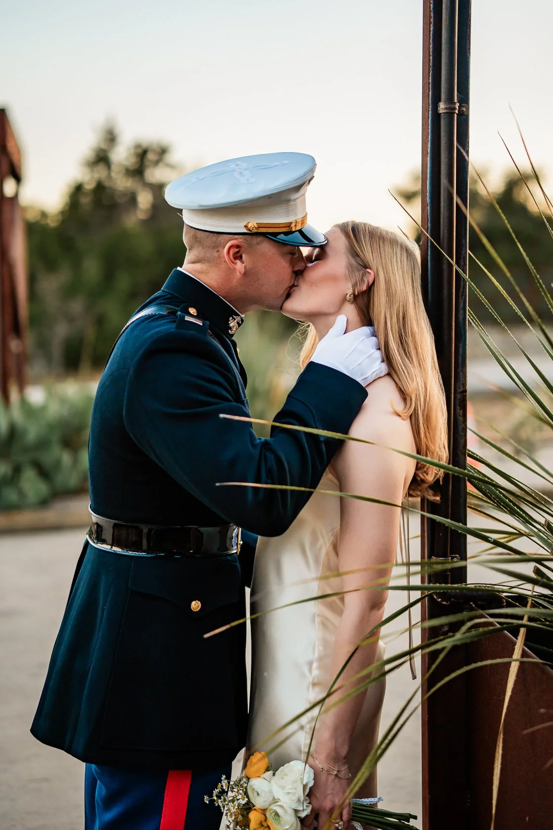 A soldier in uniform kisses a woman in a beige dress while holding a bouquet of white and yellow flowers, with a sunset sky and greenery in the background.