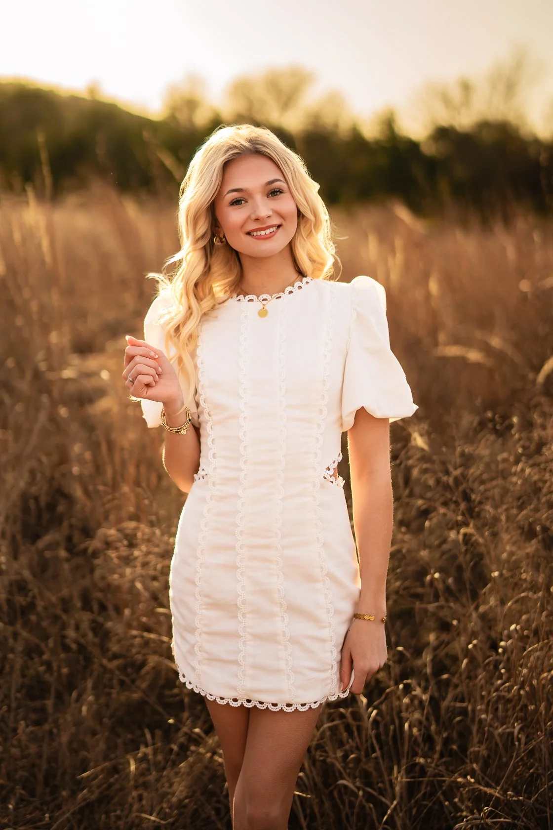 A smiling blonde woman in a white dress standing in a field at sunset.