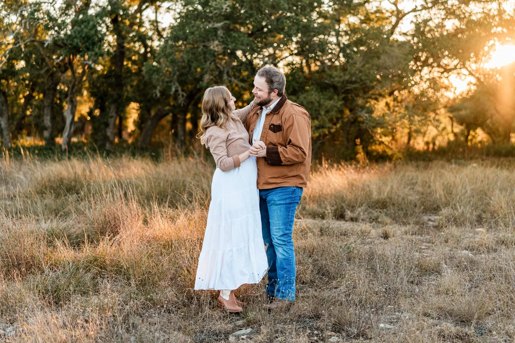 A couple dancing in a grassy field during sunset, surrounded by trees.