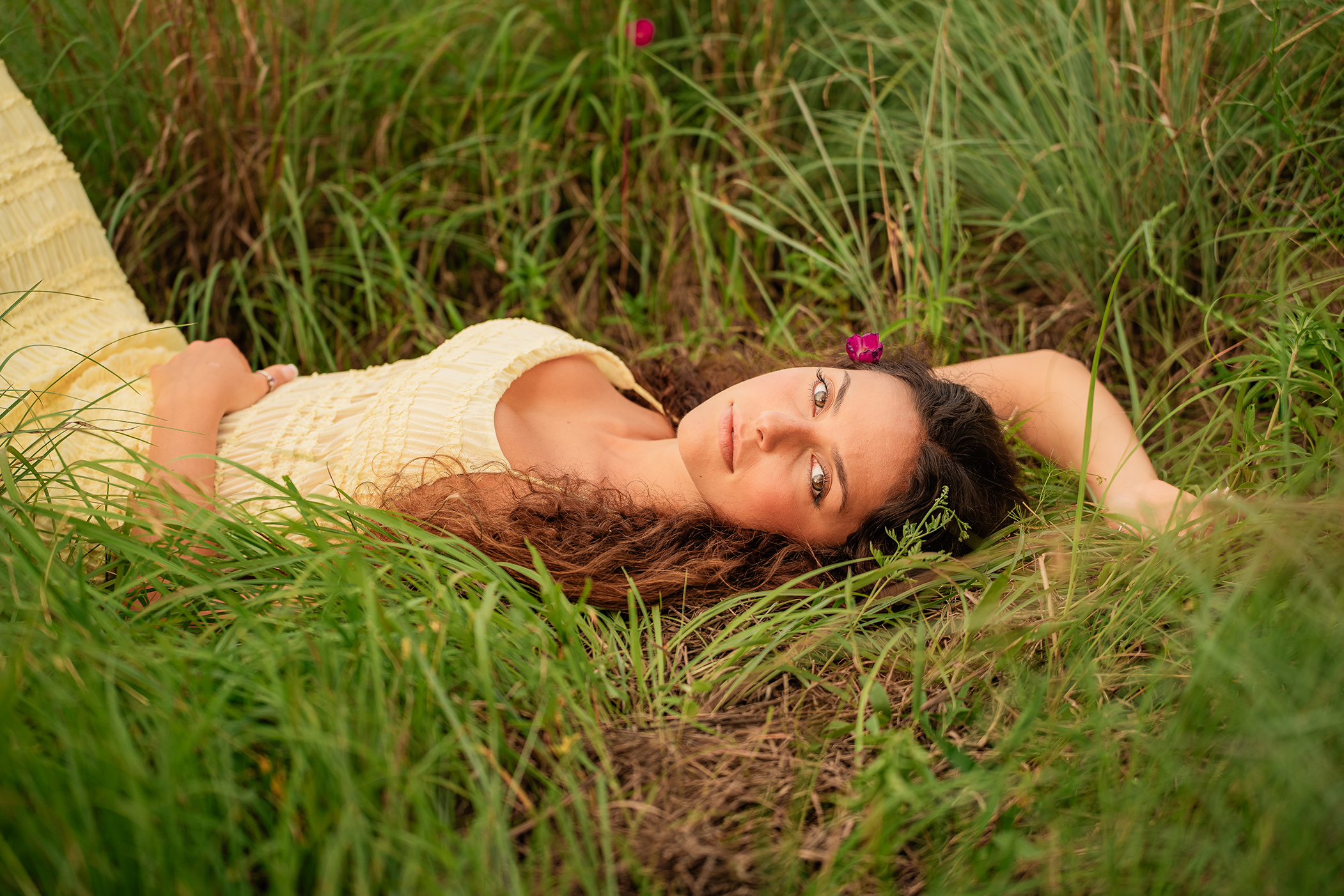 A woman with long curly hair lying on her back in tall green grass, wearing a light yellow dress with a pink flower in her hair, looking at the camera.