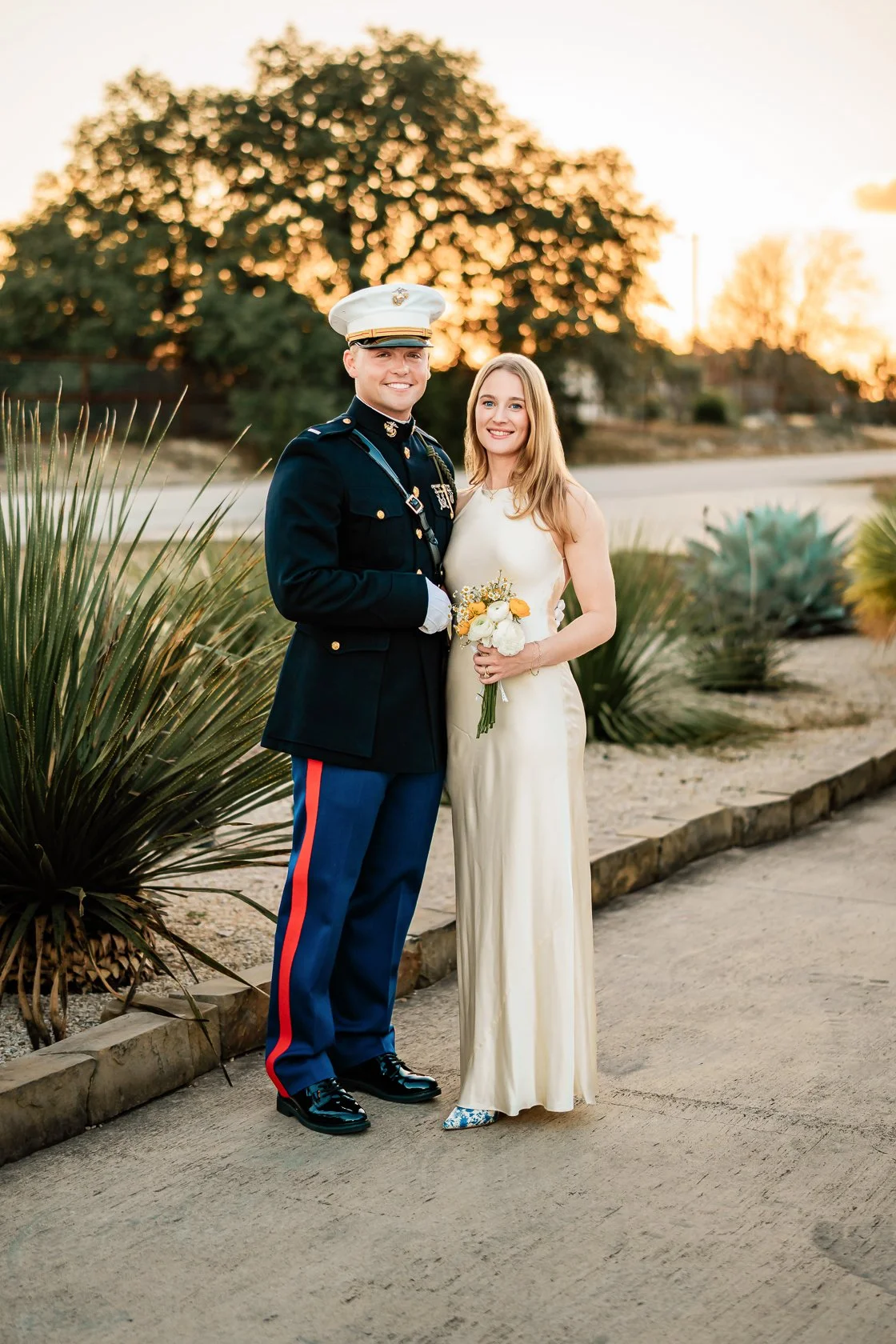 A bride and groom standing outdoors during sunset, with the groom in a formal military uniform and the bride in a white wedding dress holding a bouquet of flowers.