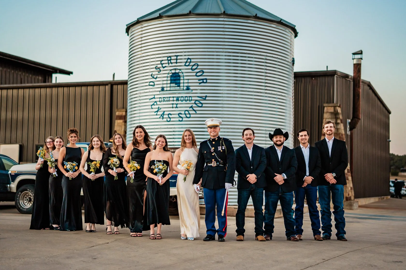 A group of people dressed in formal attire standing outside in front of a large, round metal water tank with a painted logo that says 'Desert Door, Texas, Driftwood'.