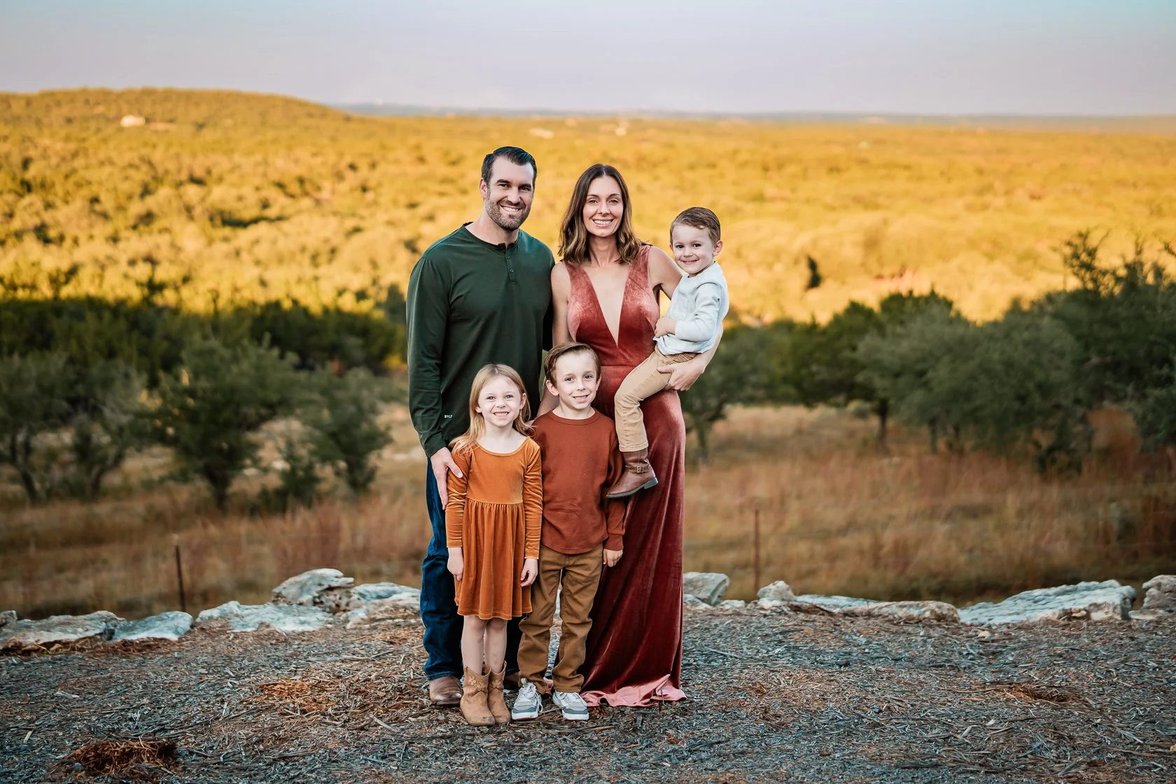 Family of five standing outdoors on rocky ground with trees and rolling hills in the background during sunset