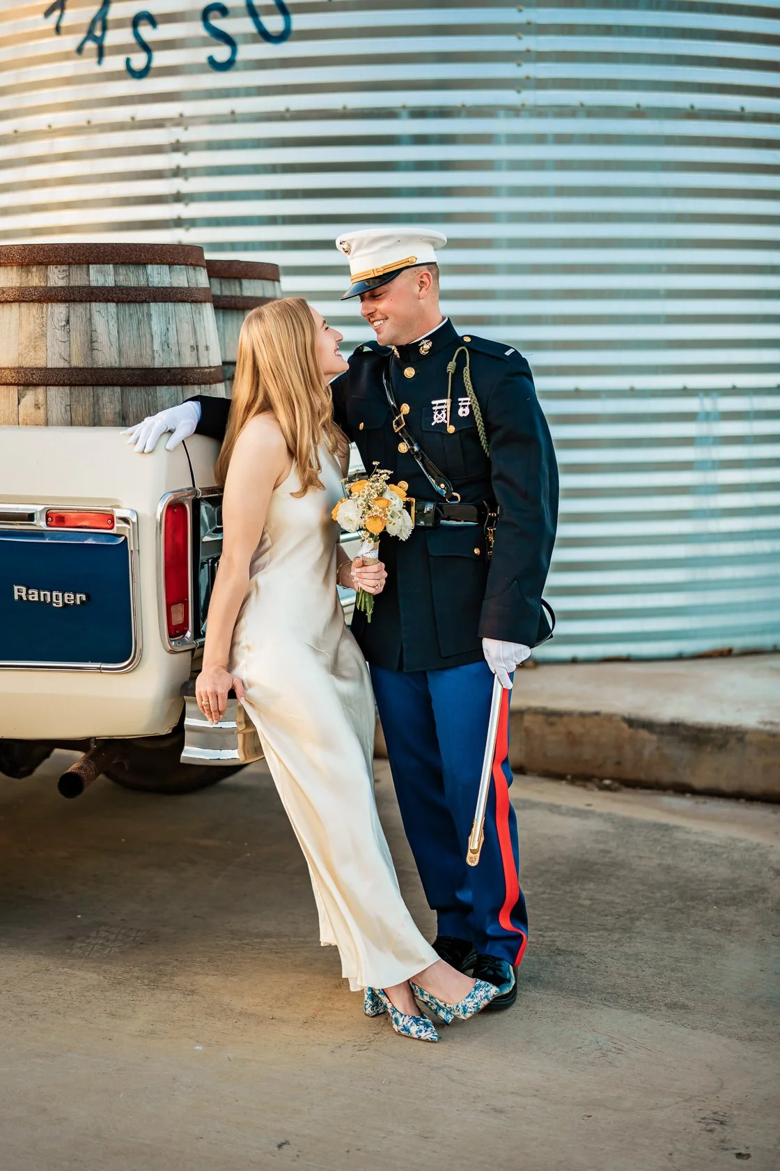 A woman in a satin wedding dress holding a bouquet of yellow and white flowers, leaning into a man in a U.S. Army dress uniform, standing beside a vintage cream-colored Ranger pickup truck, with a large industrial building in the background.