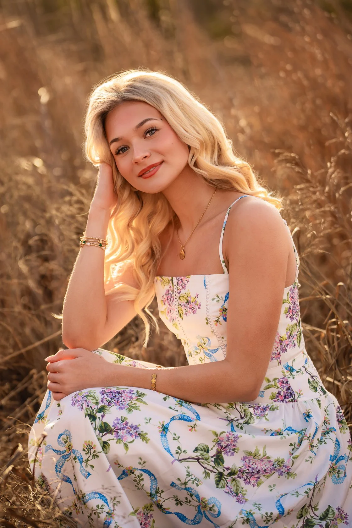 A young woman with long blonde hair sitting in a field of dry grass, wearing a white floral dress, smiling softly at the camera.