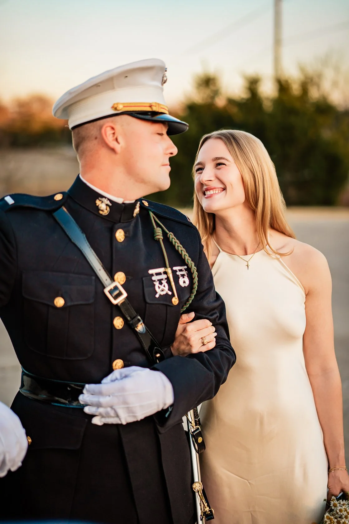 A woman in a satin dress holding hands with a uniformed military officer during sunset.