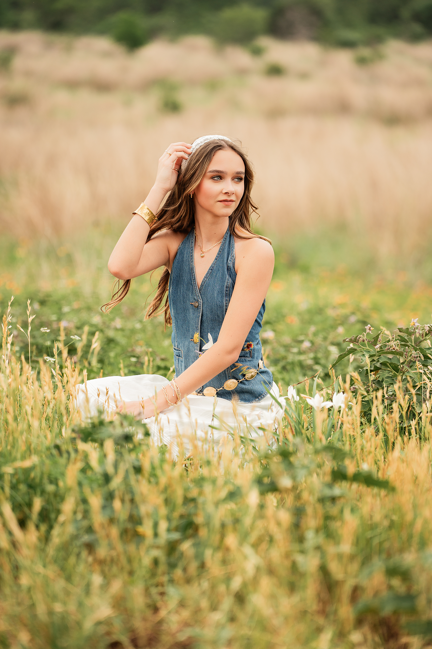 A young woman sitting in a grassy field with yellow and green plants, wearing a denim vest and white skirt, with her hair styled in loose waves and accessorized with jewelry and a headband.