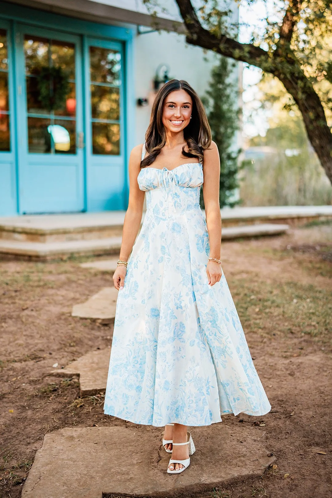 A young woman with long brown hair smiling, wearing a sleeveless white and light blue floral dress with heels, standing outdoors on a stone pathway in front of a light blue house with trees and greenery in the background.