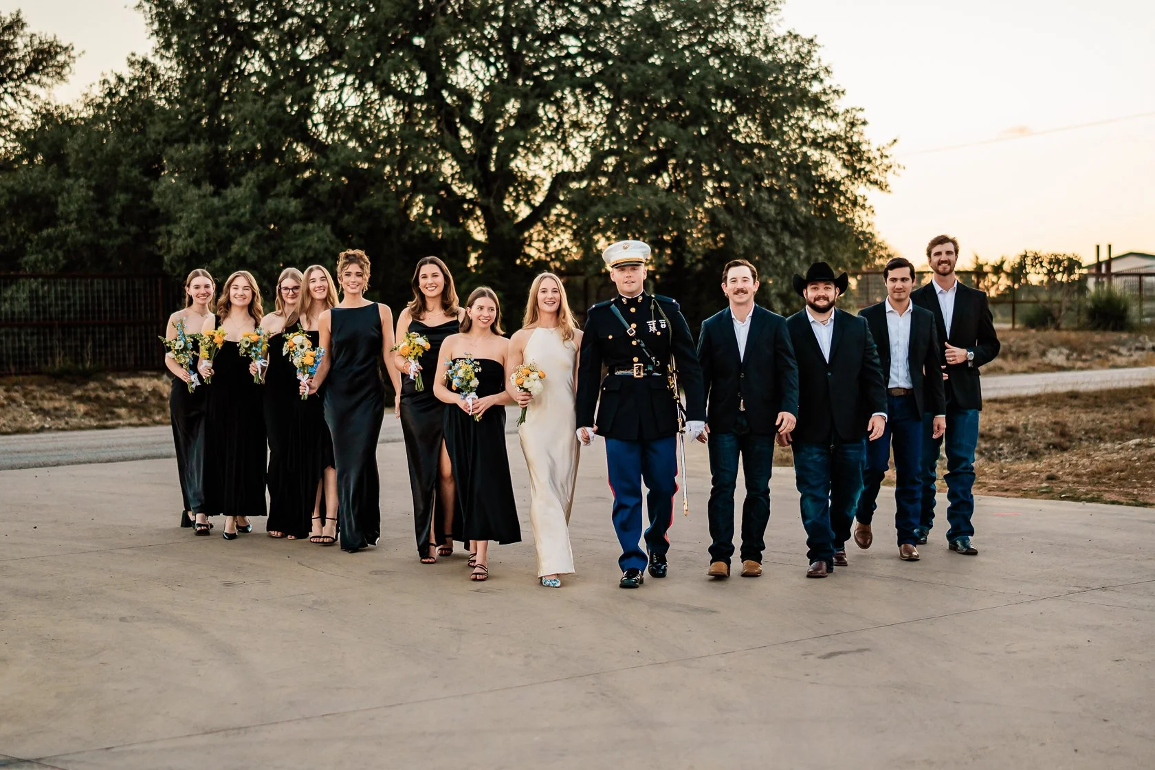 A wedding party walking outdoors on a paved path at sunset. The group includes bridesmaids in black dresses holding bouquets, a bride in a white dress, a groom in a military uniform, and several groomsmen in suits. The background features a large tre