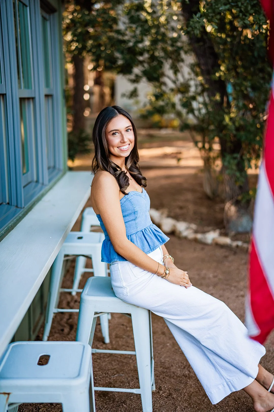 A young woman with long dark brown hair, wearing a denim strapless top and white pants, sitting on a white stool outside near a blue building and smiling at the camera.