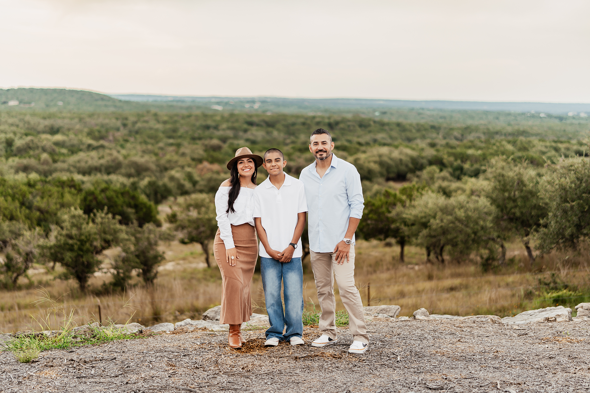 A family of three standing outdoors on a dirt path with a green landscape and trees in the background, smiling.