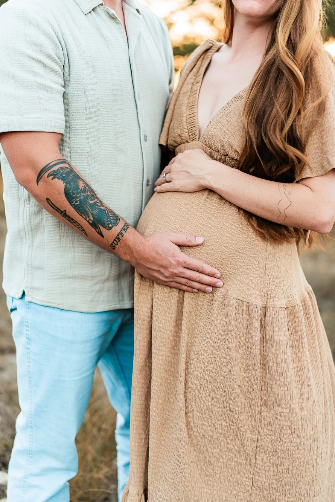 An expectant woman with long red hair, wearing a beige dress, gently holding her pregnant belly, while a person with tattooed arm and light-colored shirt places their hand on her belly during sunset outdoors.