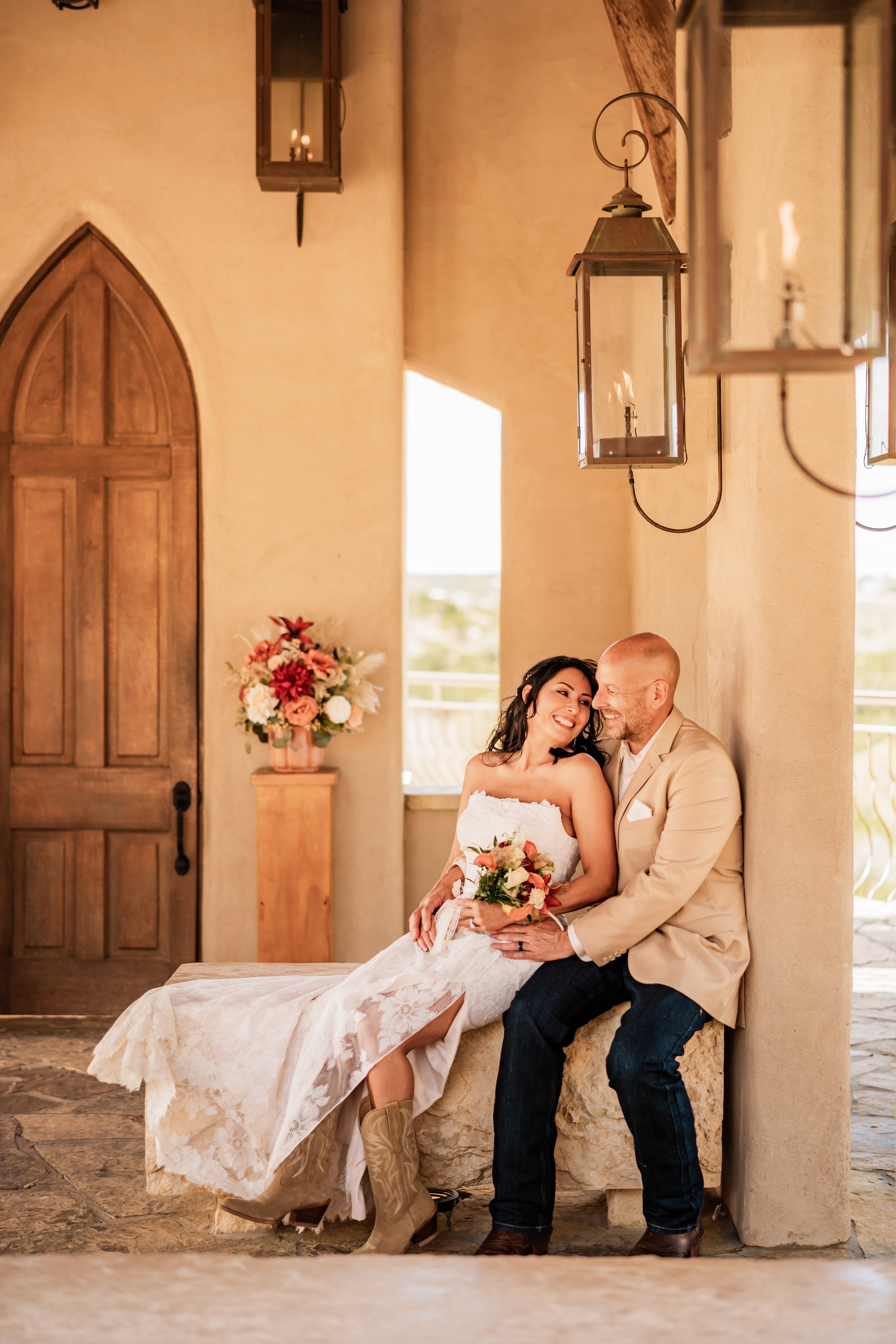 A bride and groom sitting together on a stone ledge, laughing and sharing a moment in a warmly lit, rustic setting with hanging lanterns and a floral arrangement nearby.