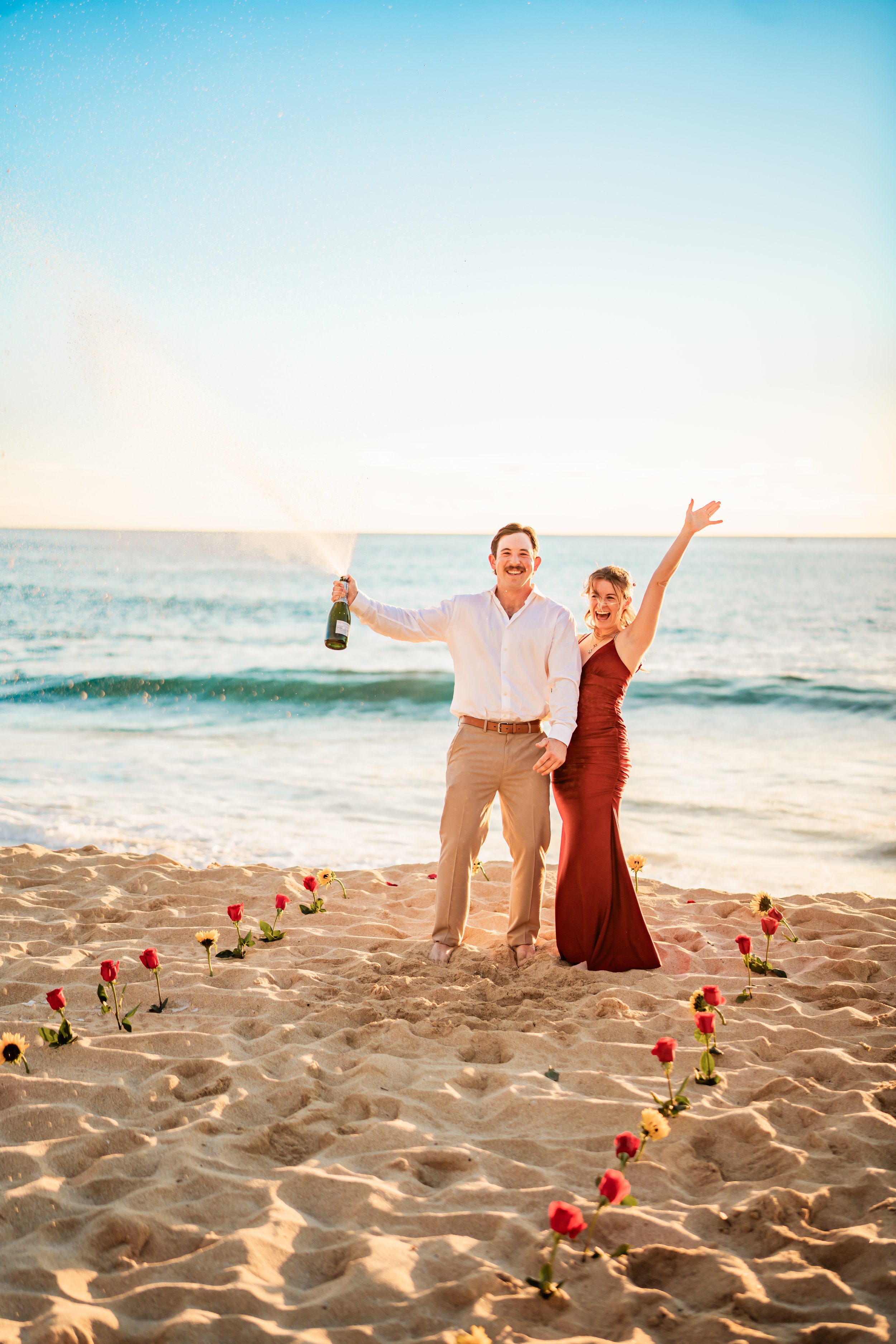 A joyful couple celebrating on the beach, with the man holding a champagne bottle and the woman raising her arm, surrounded by a trail of colorful flowers in the sand, with ocean waves and a clear sky in the background.