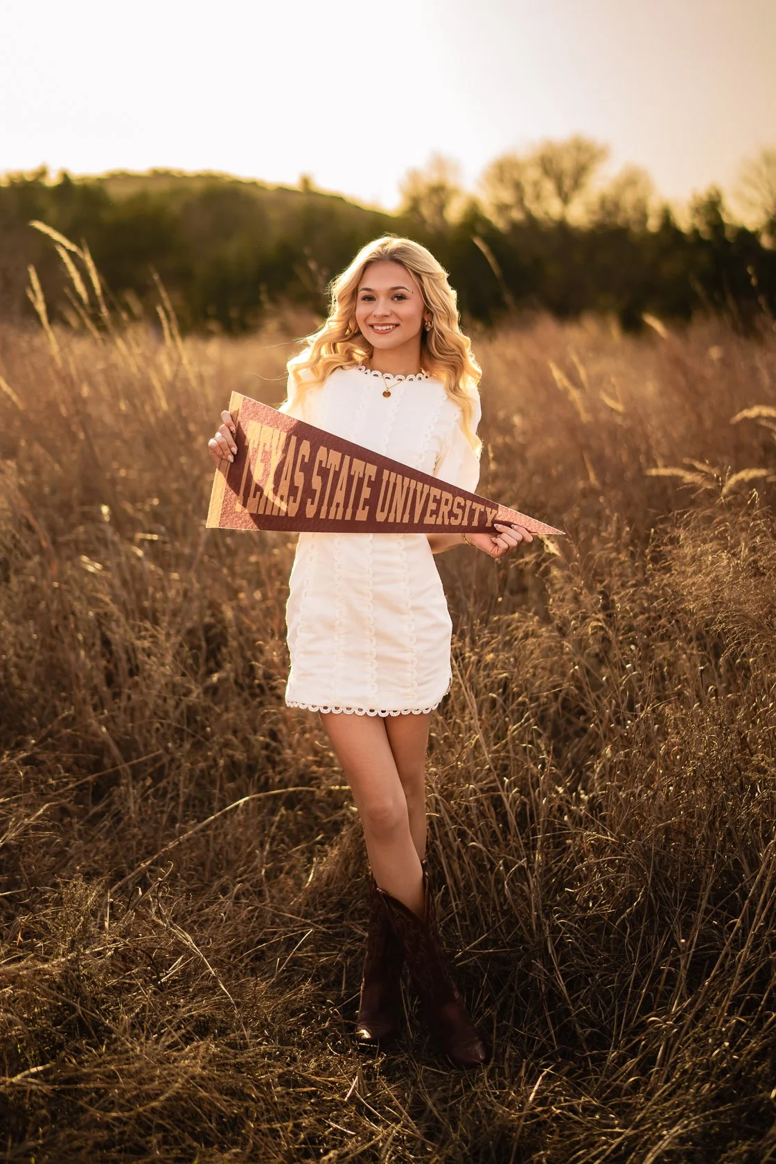 A young woman with blonde hair standing in a field of tall grass at sunset, holding a maroon and gold pennant that reads 'TEXAS STATE UNIVERSITY', smiling at the camera.