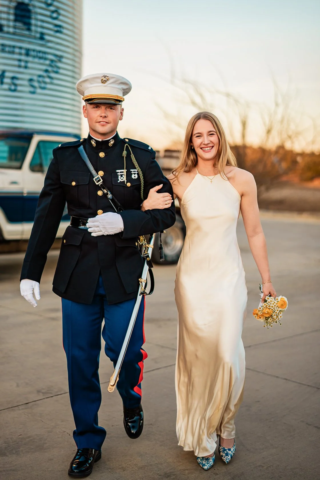 A man in a military dress uniform and a woman in a light-colored satin dress walk together outdoors during sunset, with the woman holding a small bouquet of flowers.