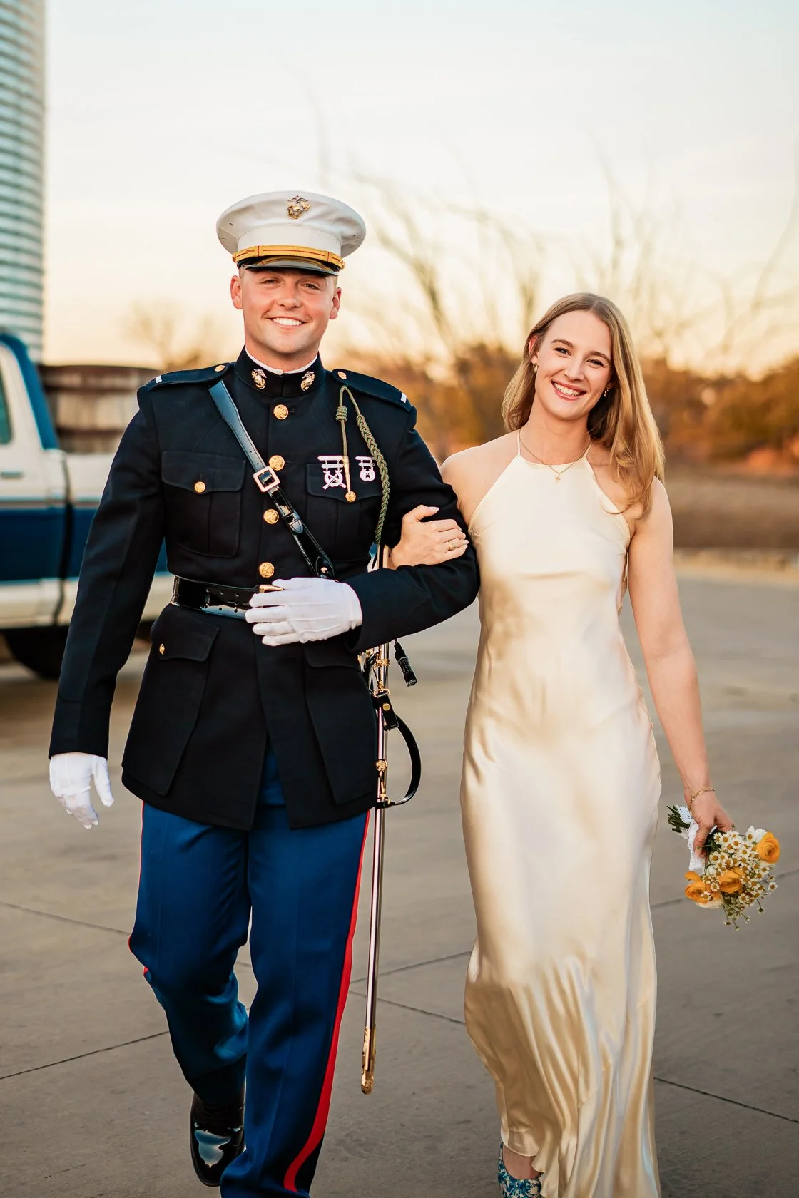 A young man in a United States Marine Corps uniform walking arm-in-arm with a young woman in a cream-colored dress, holding a small bouquet of flowers, outside during sunset.