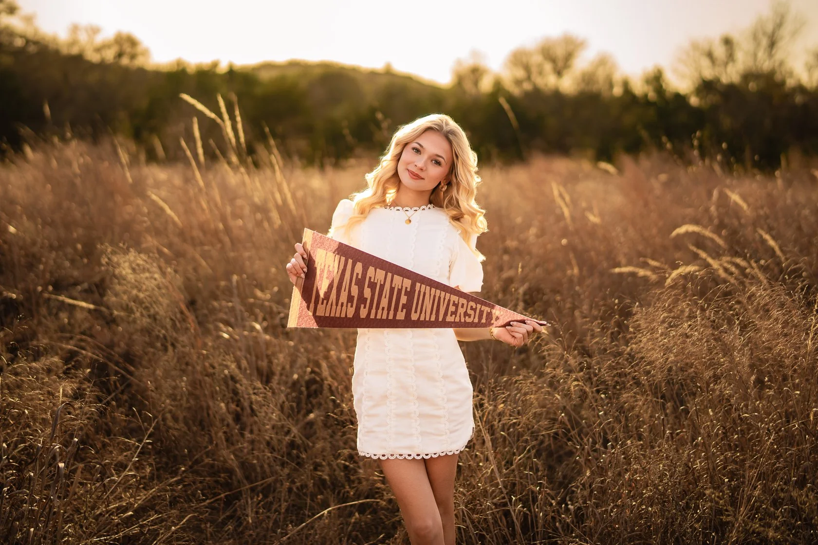 A young woman with blonde curly hair holding a 'Texas State University' pennant, standing in a field of tall, dry grass during sunset.