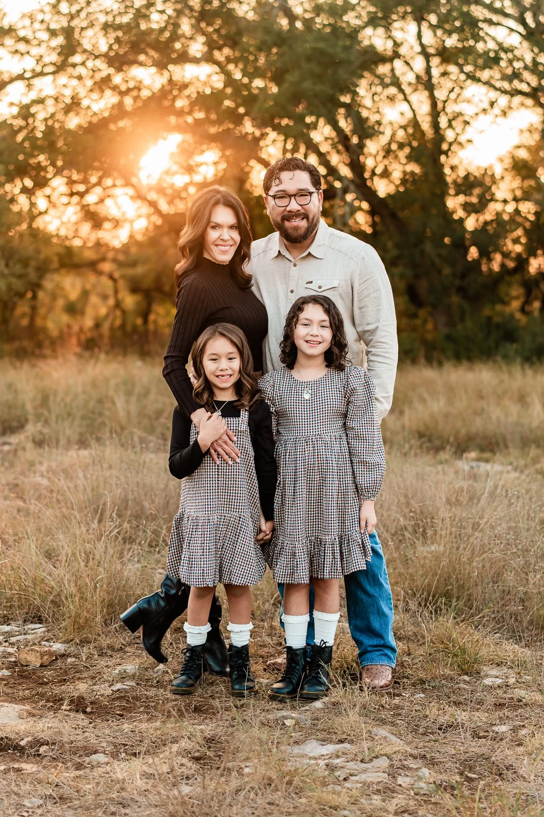 A family of four standing outdoors in a field with trees at sunset. The mother and father are smiling behind their two daughters, who are holding hands and wearing matching dresses. The background has warm, golden sunlight filtering through the trees