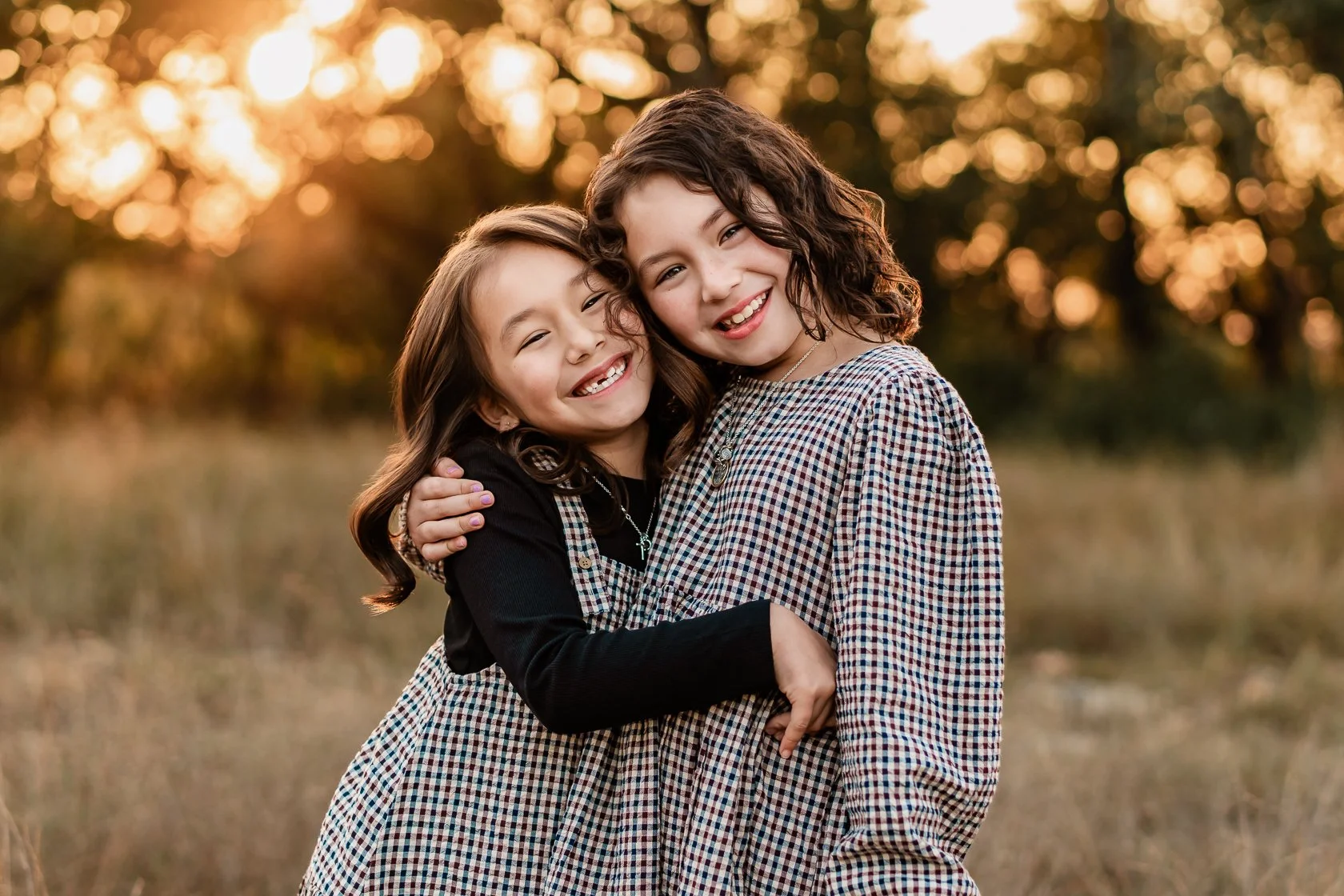Two young girls hugging and smiling outdoors during sunset, with trees and warm sunlight in the background.