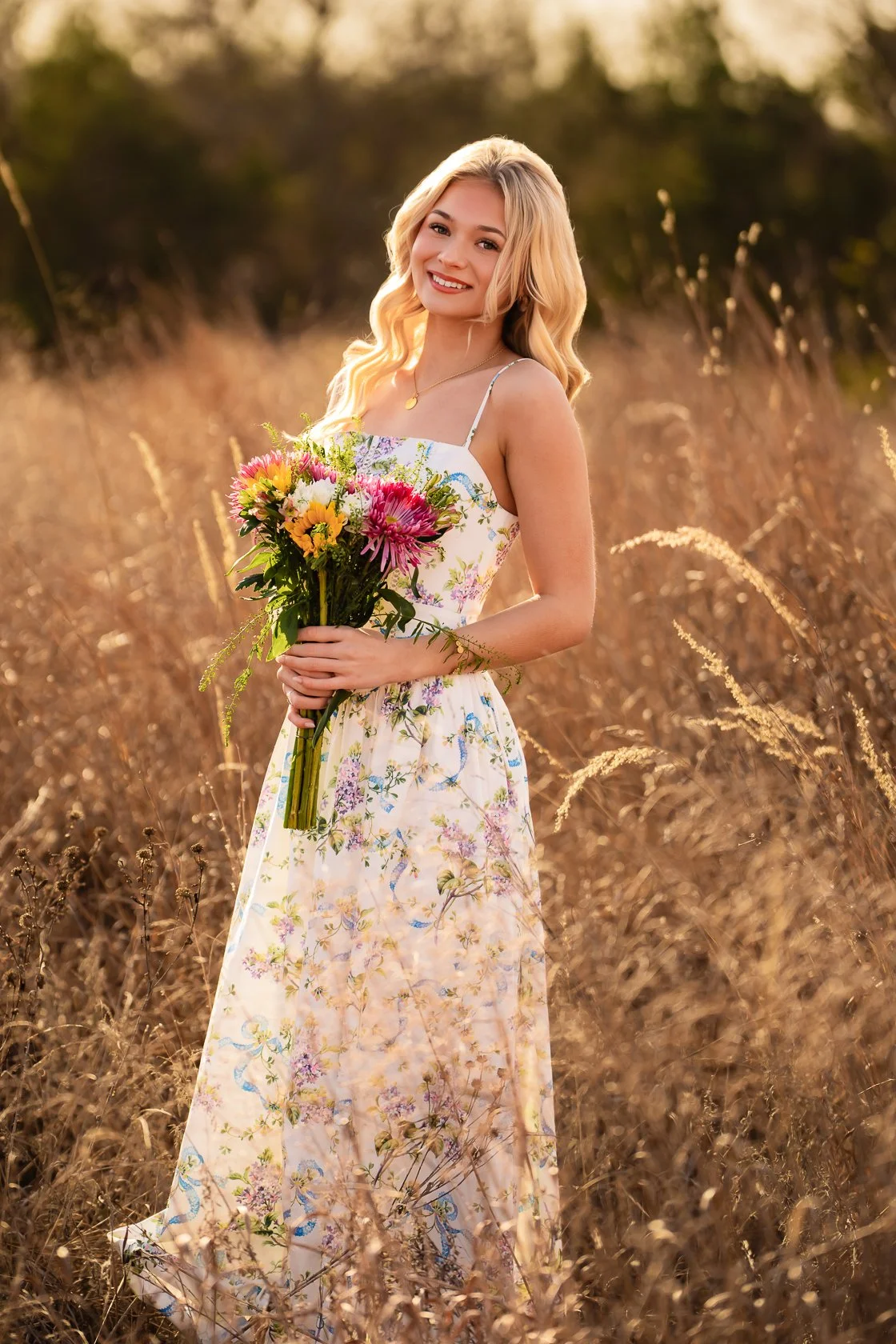 A young woman with blonde hair smiling and holding a bouquet of colorful flowers in a field of tall, dry grass during sunset.