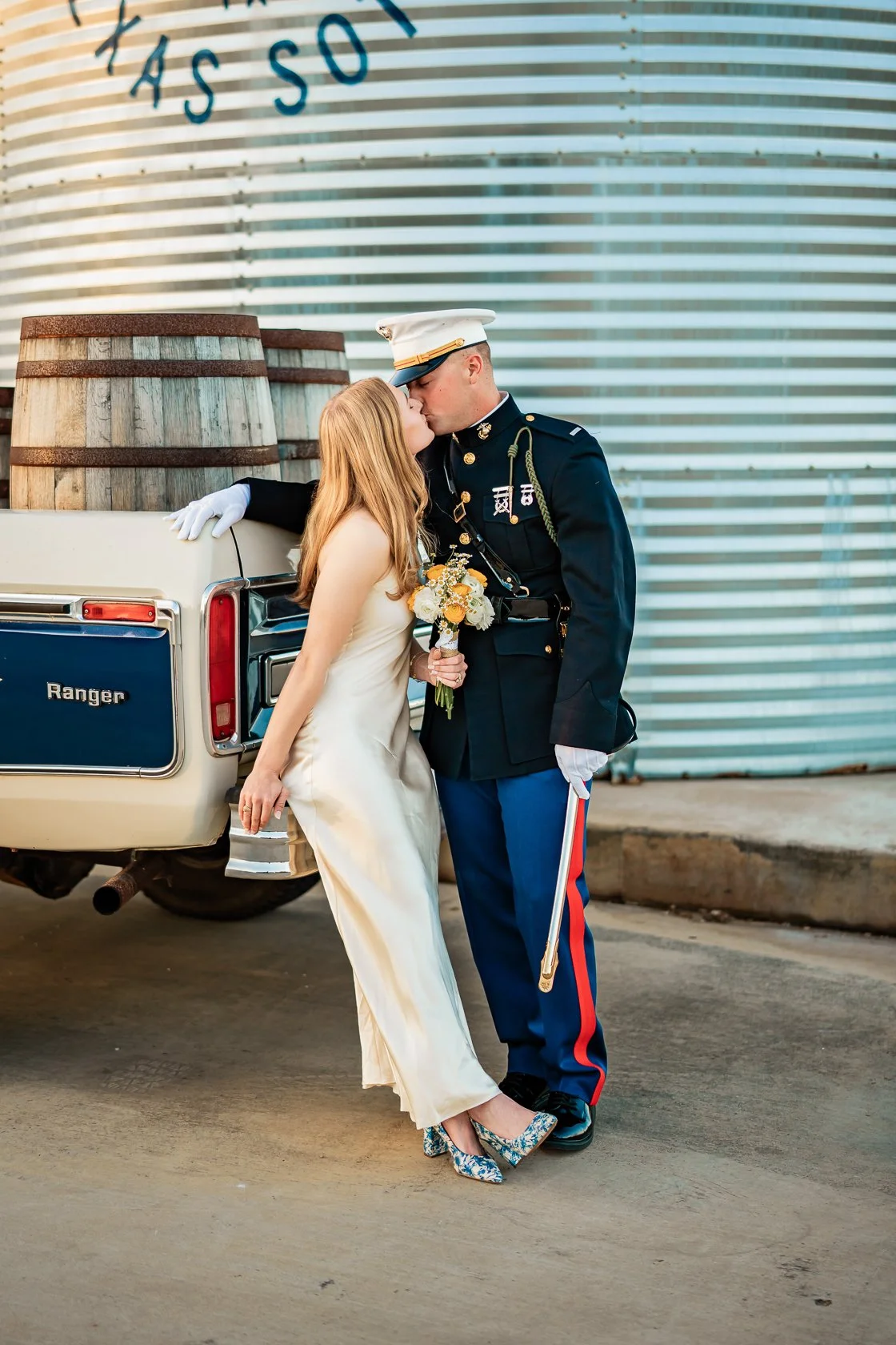 A bride and a soldier share a kiss in front of a vintage pickup truck with wooden barrels on top, in an industrial setting.