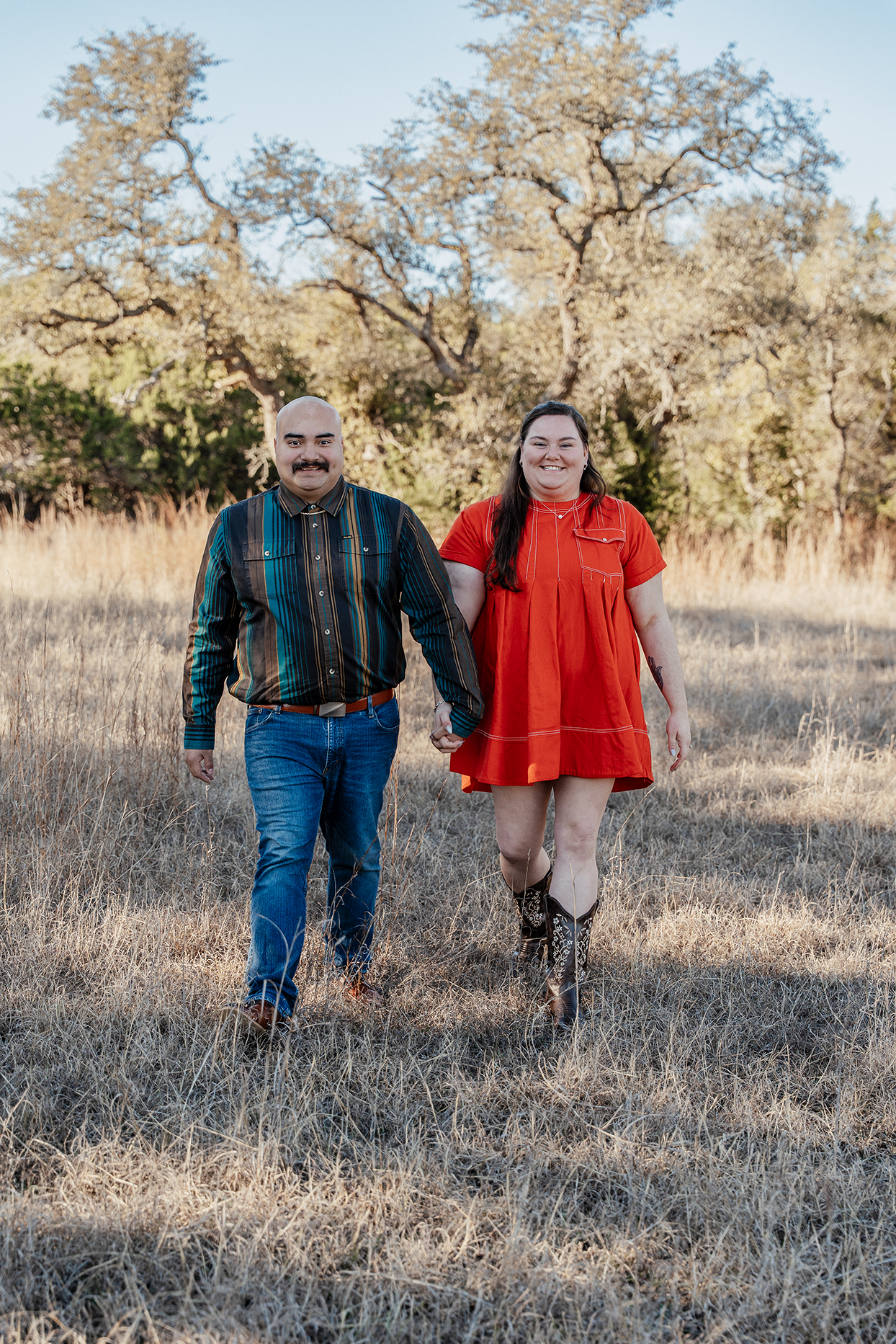 A couple walking hand in hand through a grassy field with trees in the background, smiling at the camera.
