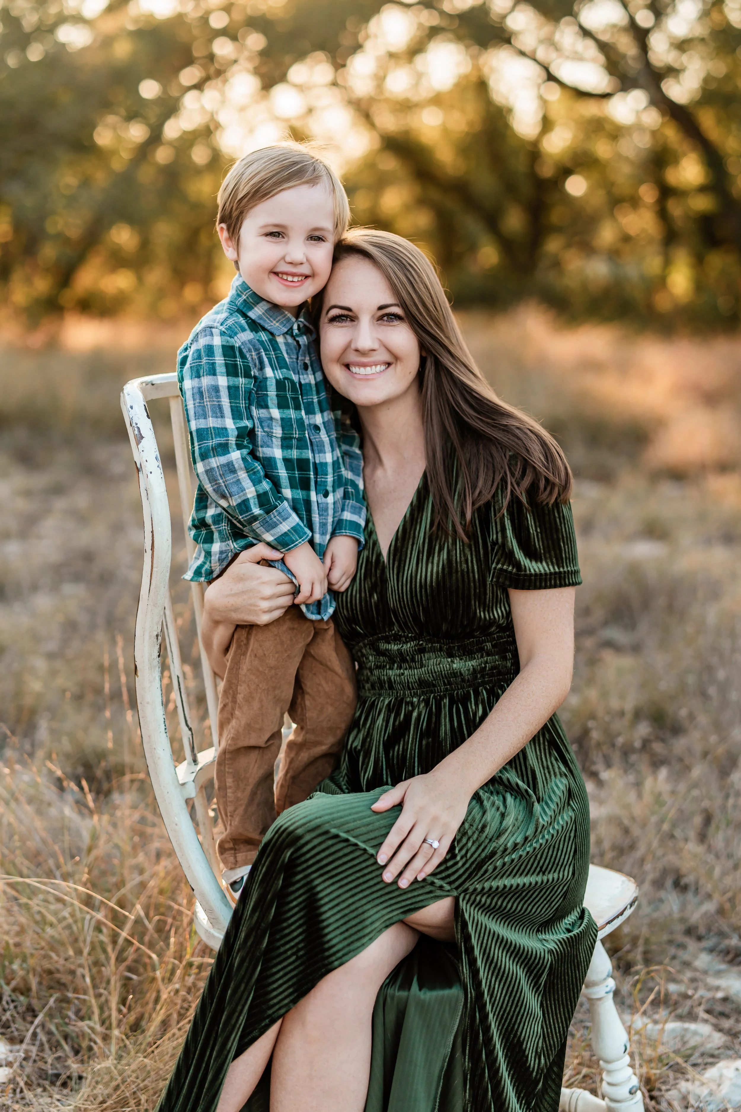 A woman and a young boy sitting outdoors on a white chair during sunset, smiling at the camera.