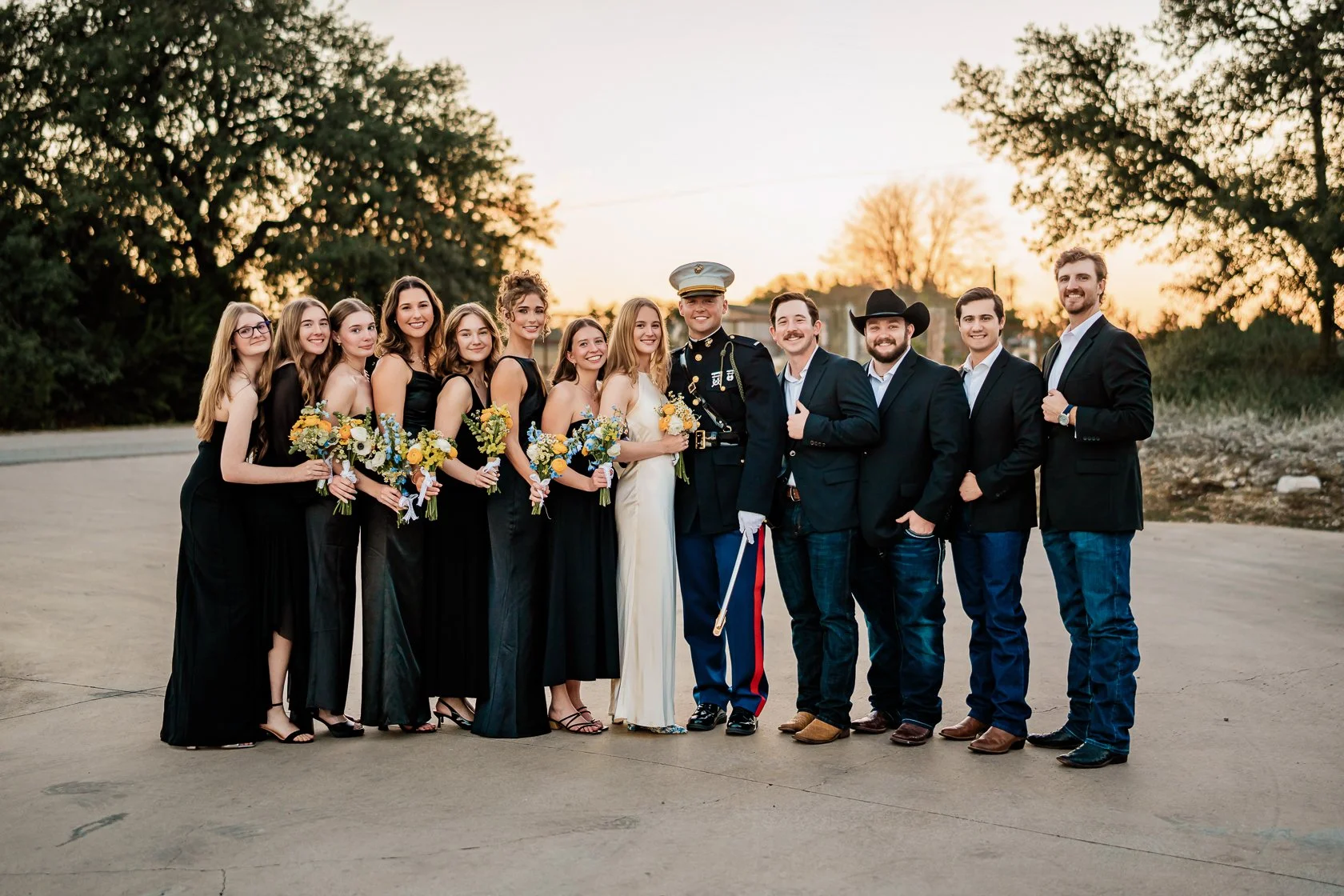 Group of people at a wedding, including bridesmaids, groomsmen, and the bride and groom, standing outdoors during sunset.
