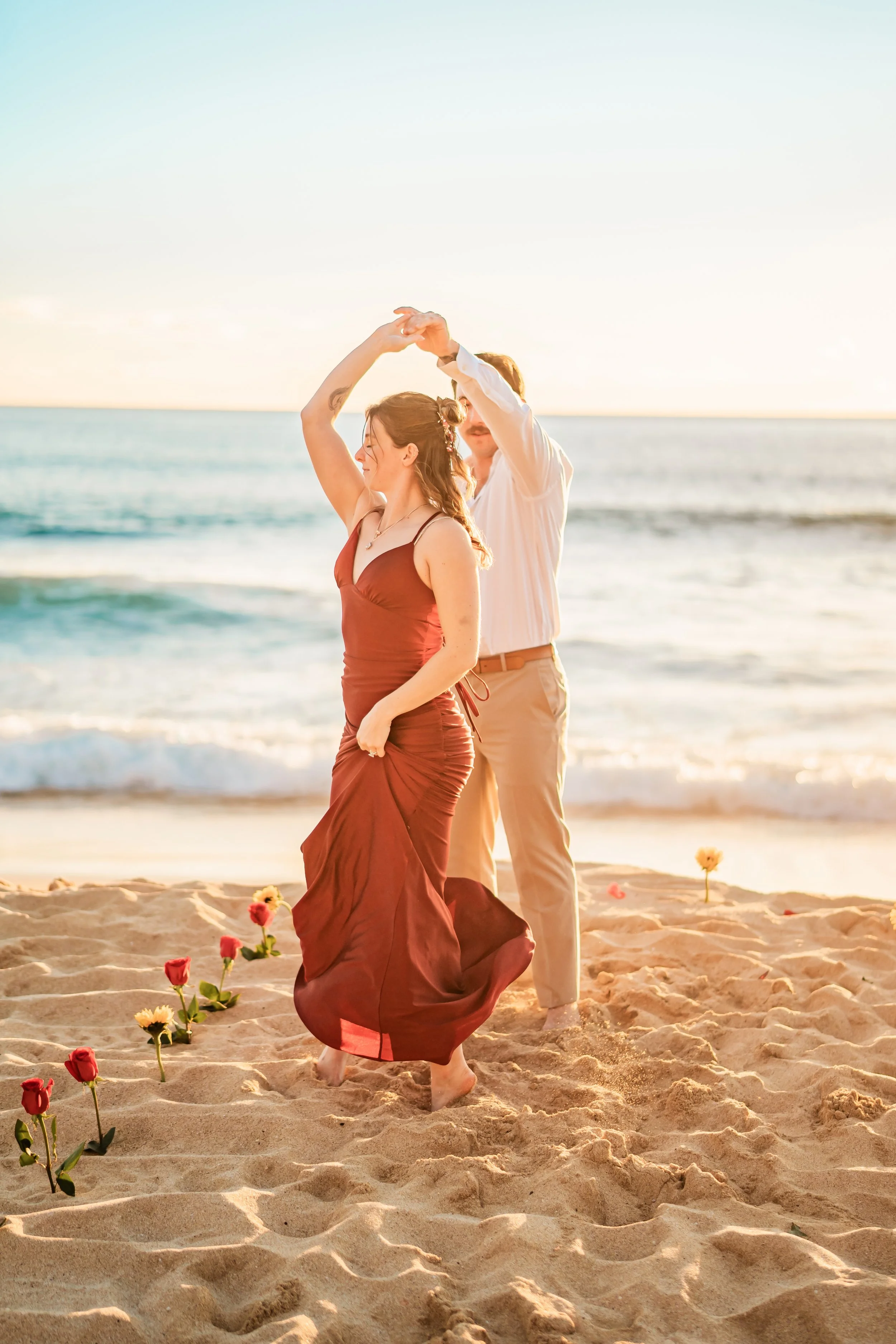 A couple dancing barefoot on a sandy beach at sunset, with flowers planted in the sand and ocean waves in the background.