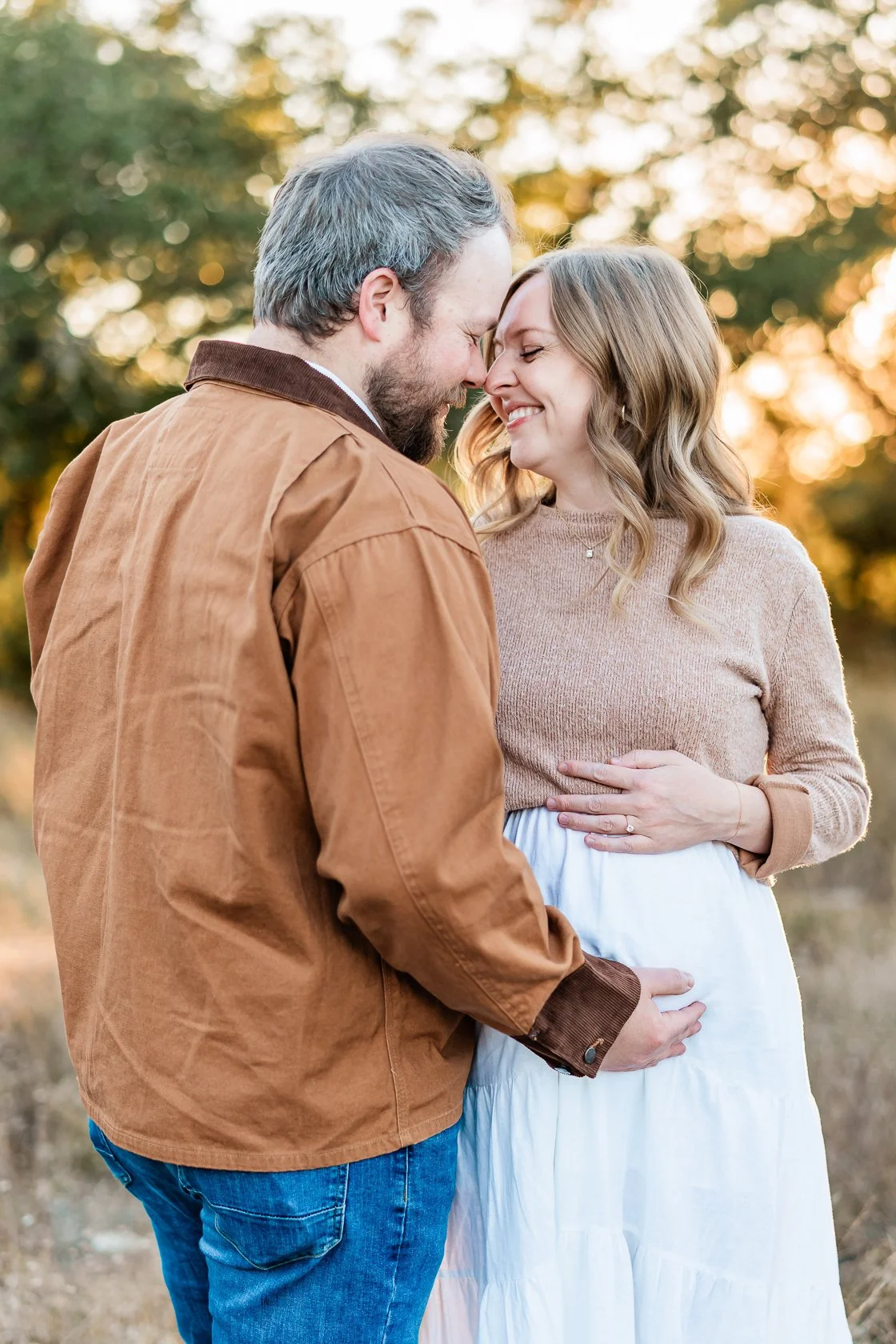 A couple faces each other with their foreheads touching, smiling, outdoors during sunset, with trees in the background.