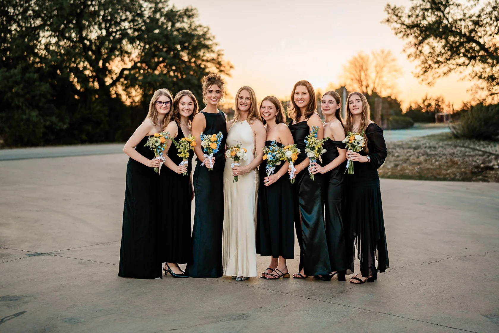 Group of women dressed in formal evening gowns holding bouquets during sunset outdoors.