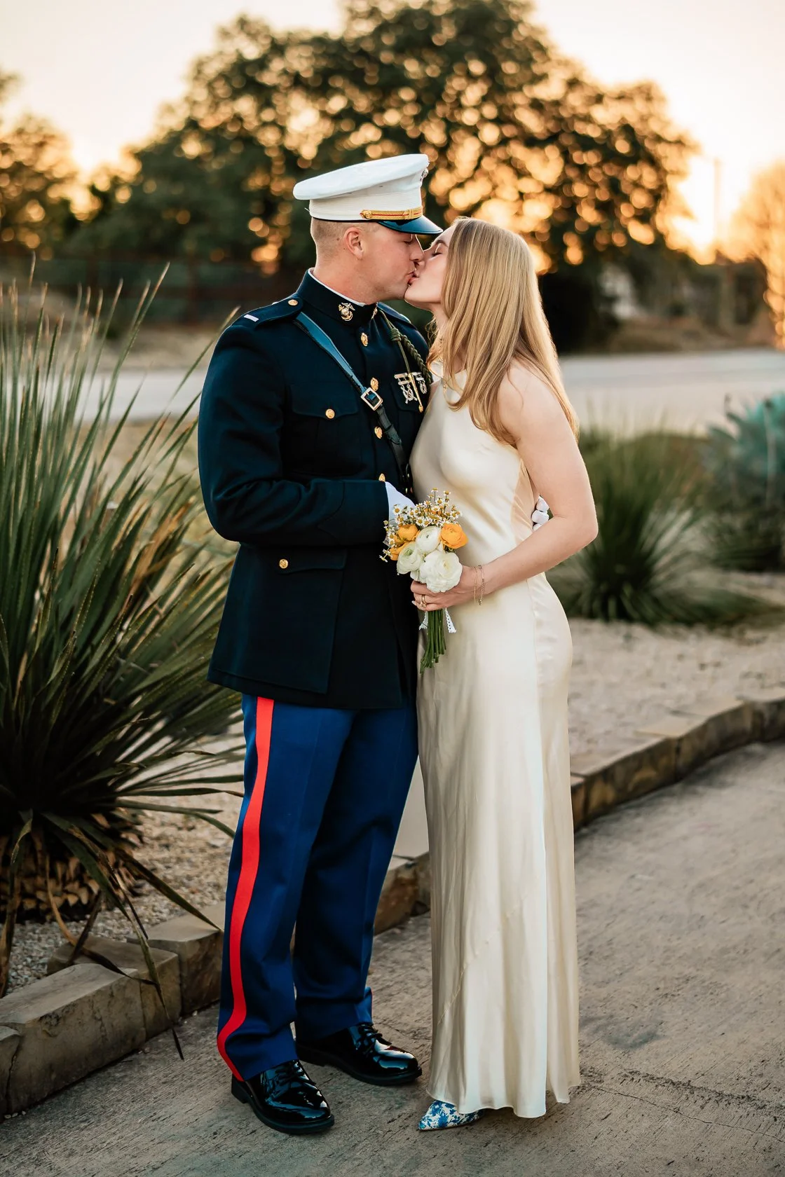 A couple sharing a kiss during their wedding; the man is in a U.S. military uniform and the woman is in a white satin dress holding a bouquet of flowers, outdoors at sunset.