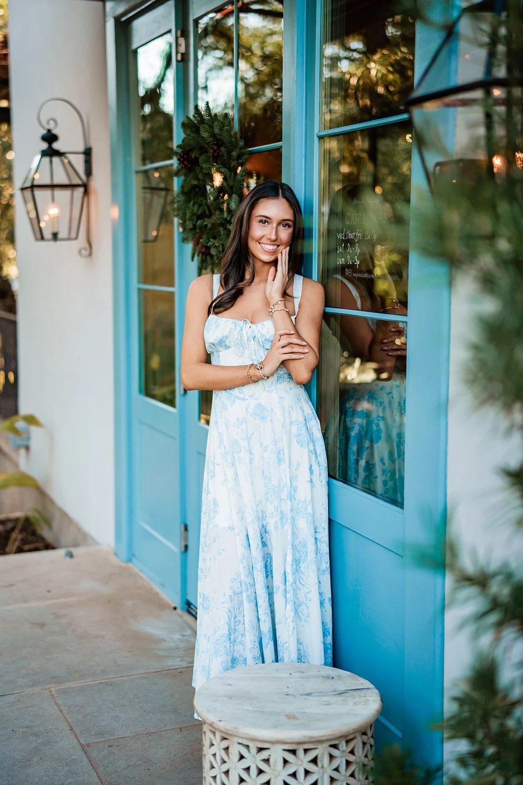 A smiling woman in a white and blue floral maxi dress stands outside a blue door with glass panels, touching her face with her right hand. She is decorated with bracelets and has long dark hair. The setting appears to be outdoors with greenery and a 