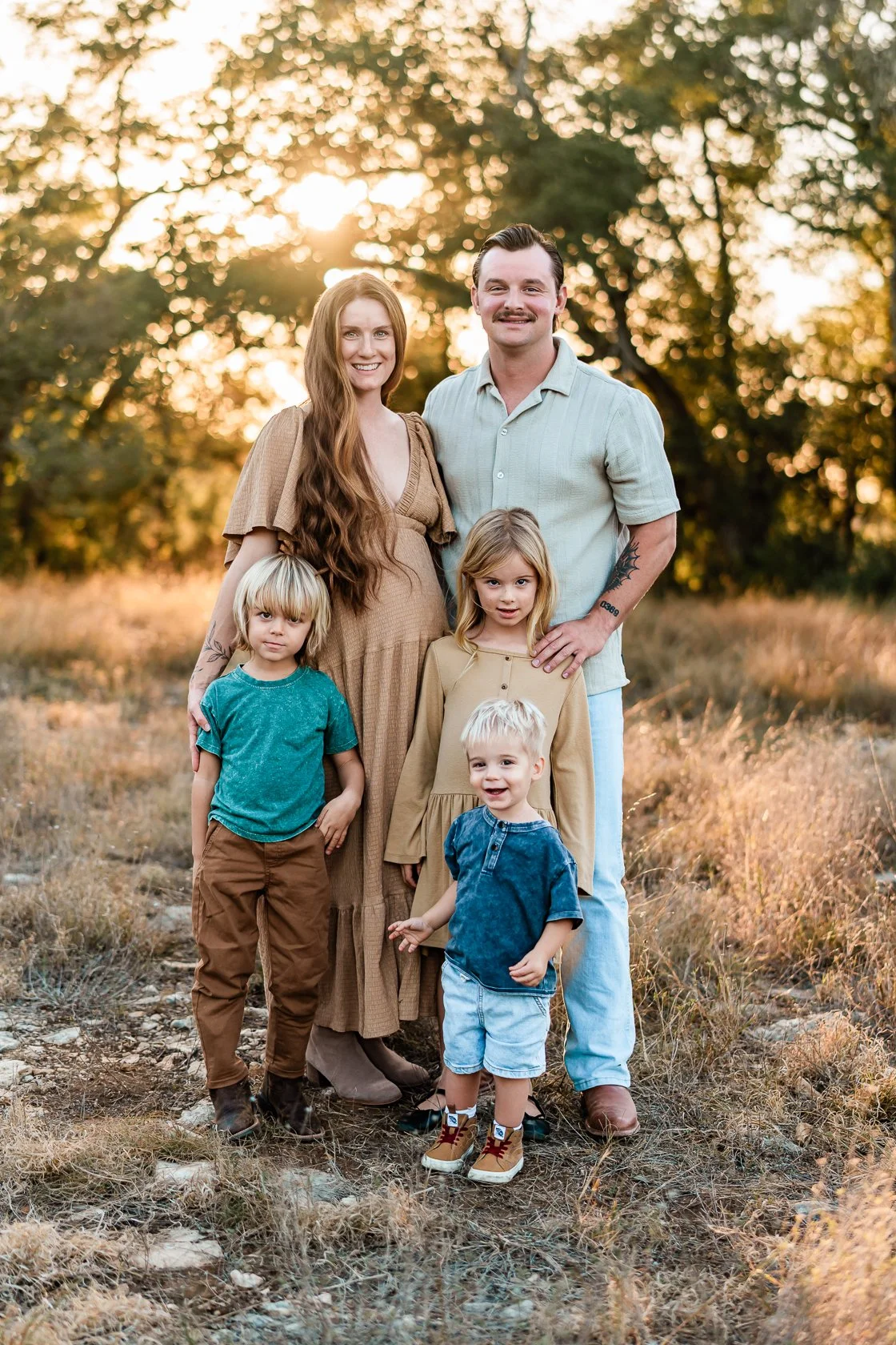 A family of six posing outdoors during sunset, with trees and golden light in the background.
