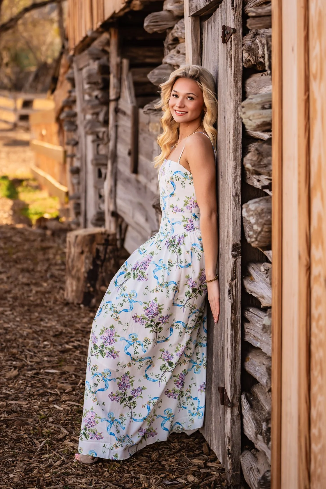 Young woman in a floral dress leaning against a rustic wooden shed outdoors during sunset.
