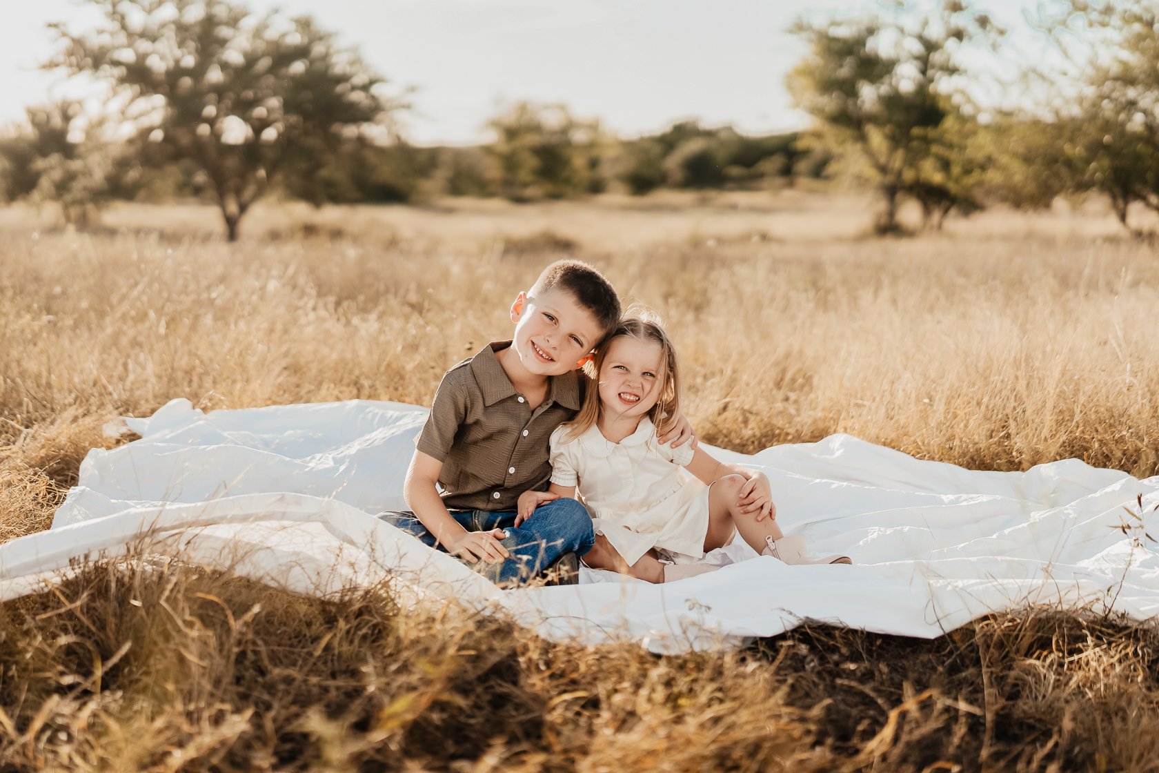 Two young children, a boy and a girl, sitting on a white blanket in a grassy field with trees in the background, smiling and enjoying a sunny day.
