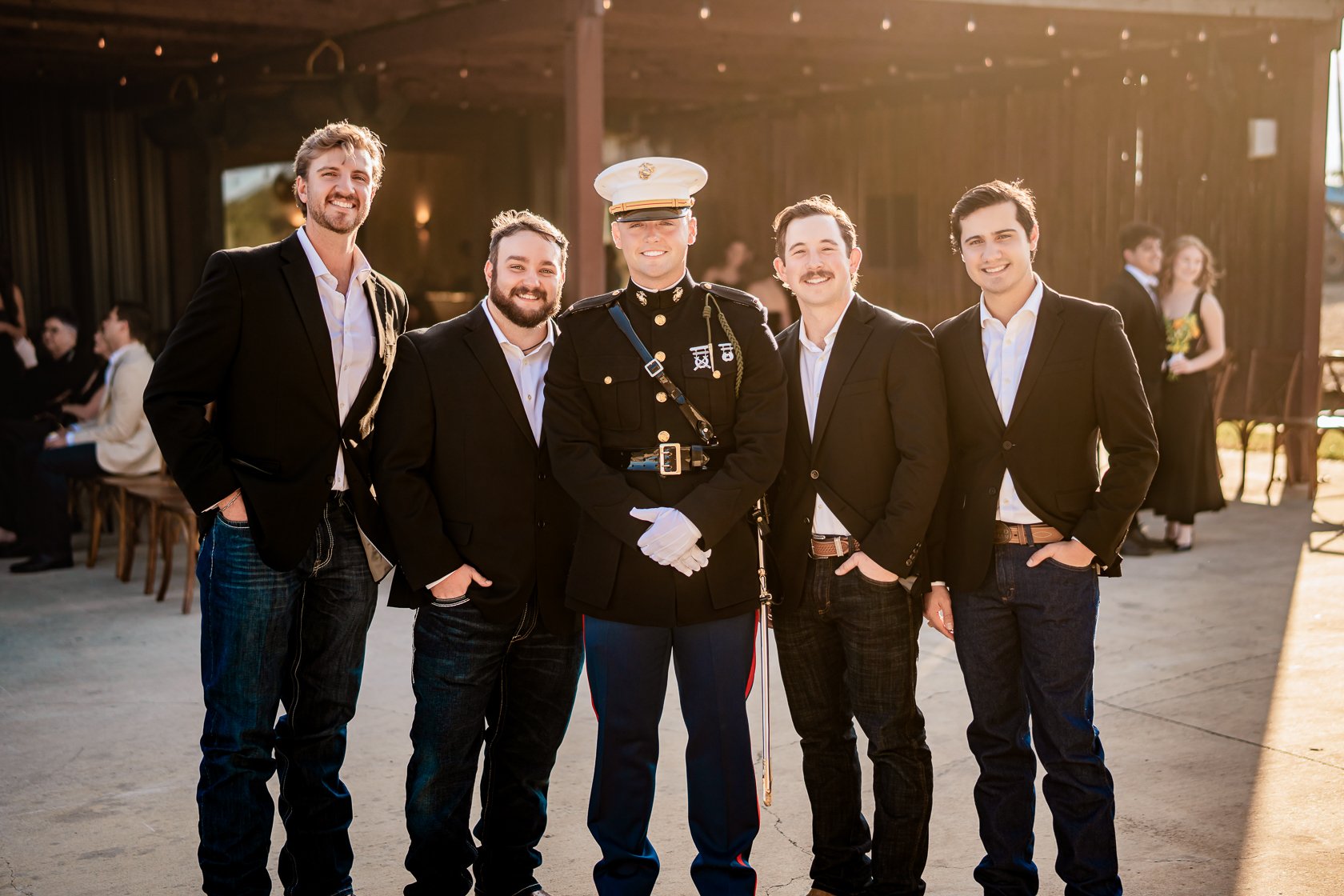 Group of five men, one in military uniform, standing and smiling at a social gathering outdoors during the daytime.