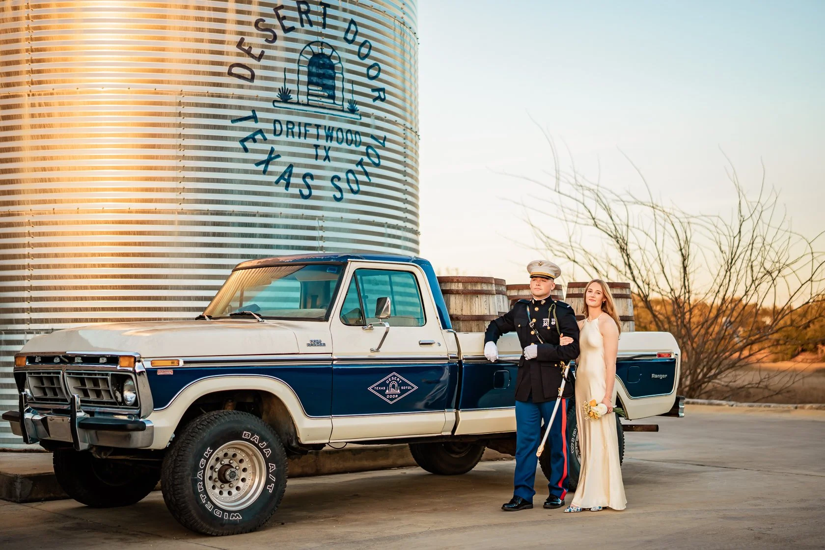 A bride and groom standing outside near a vintage pickup truck, with a large water tank in the background displaying the Texas flag and a logo for Driftwood Texas. The groom is dressed in a military uniform, and the bride is wearing a long white wedd