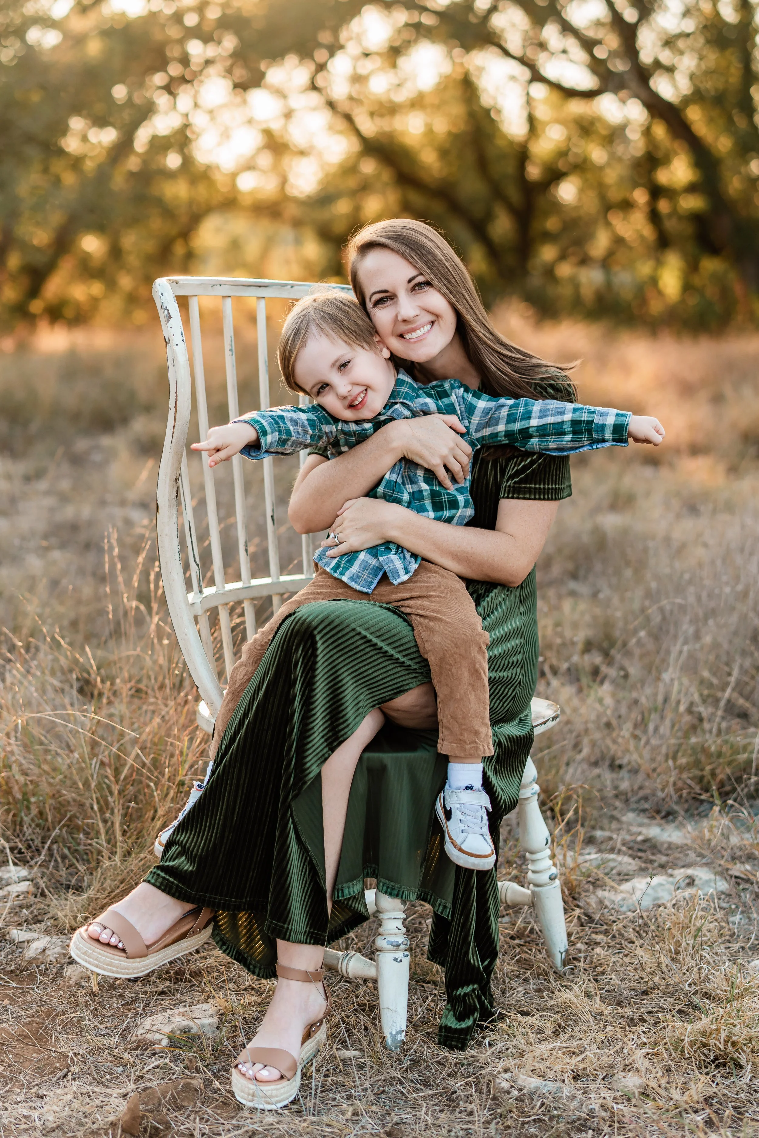 A woman and a young boy sitting on an old white wooden chair outdoors during sunset, smiling and embracing with trees and golden light in the background.