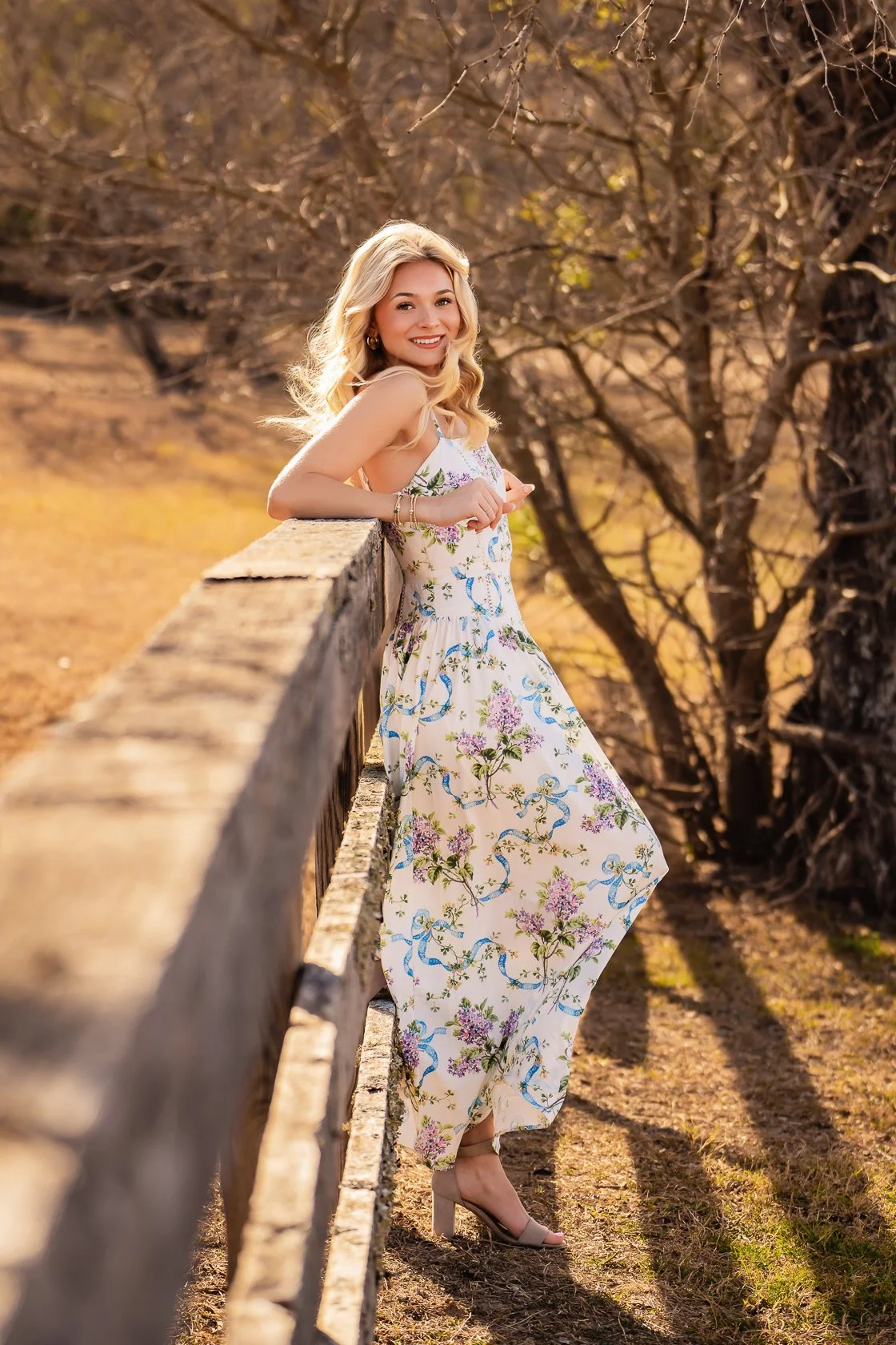 A young woman with blonde hair wearing a floral dress stands beside a wooden fence in a natural outdoor setting, smiling at the camera during golden hour.