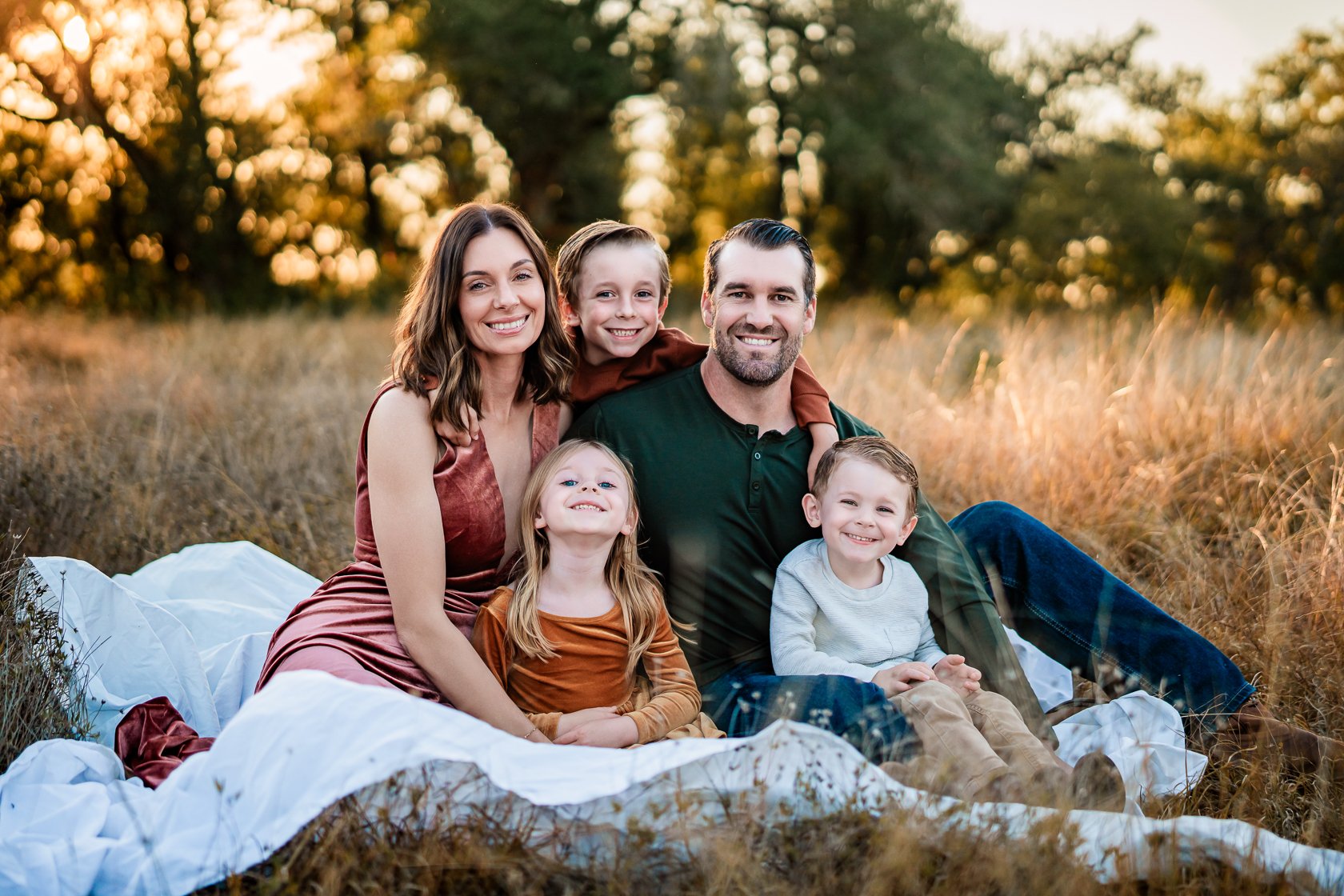 A smiling family of six sitting on a white blanket in a field during sunset, surrounded by tall grass and trees.