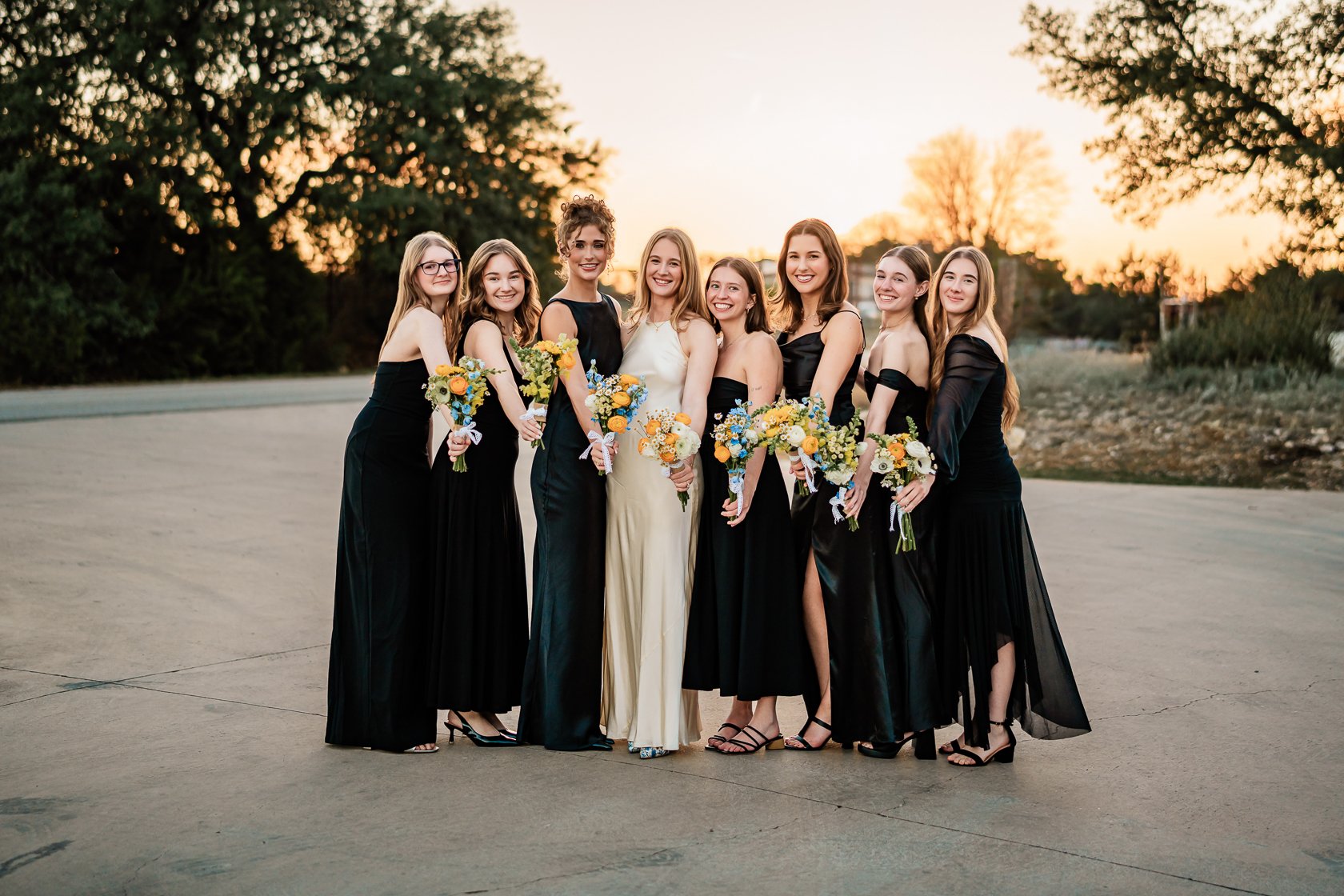 A group of nine women dressed in formal gowns standing together outdoors at sunset, holding bouquets of flowers.