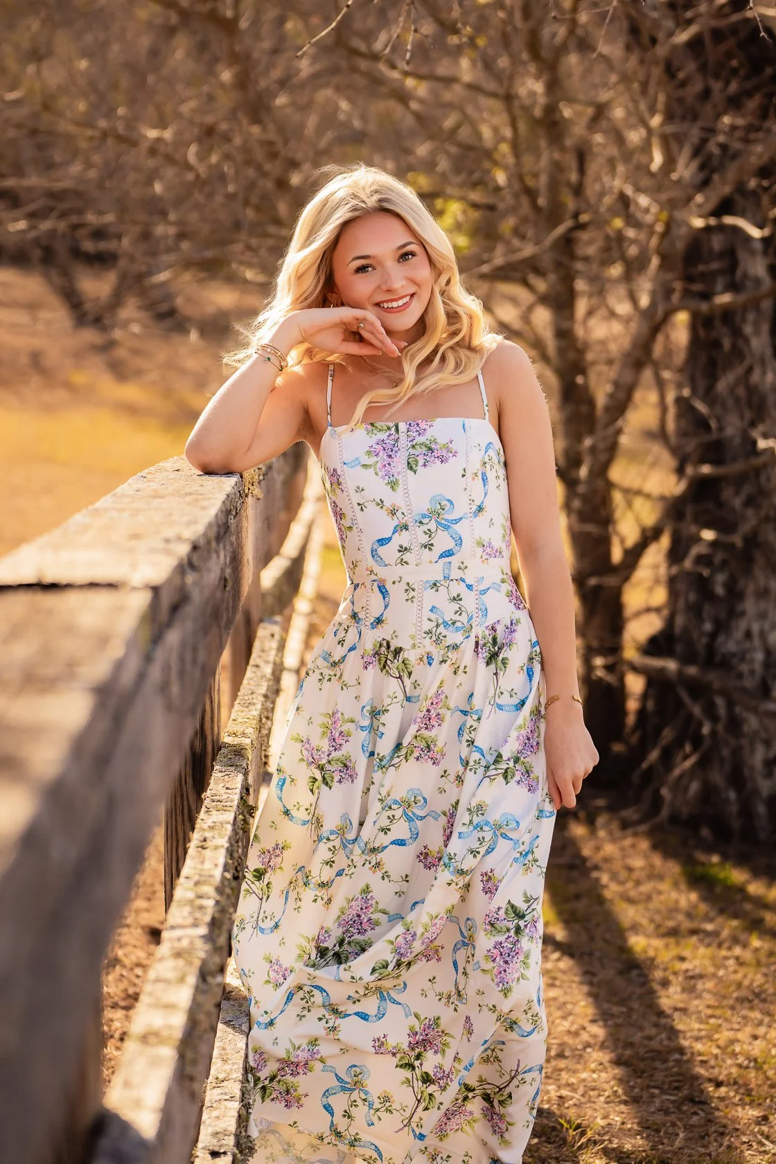 A young woman with blonde curly hair wearing a white floral and snake pattern dress, leaning on a wooden railing outdoors during golden hour.